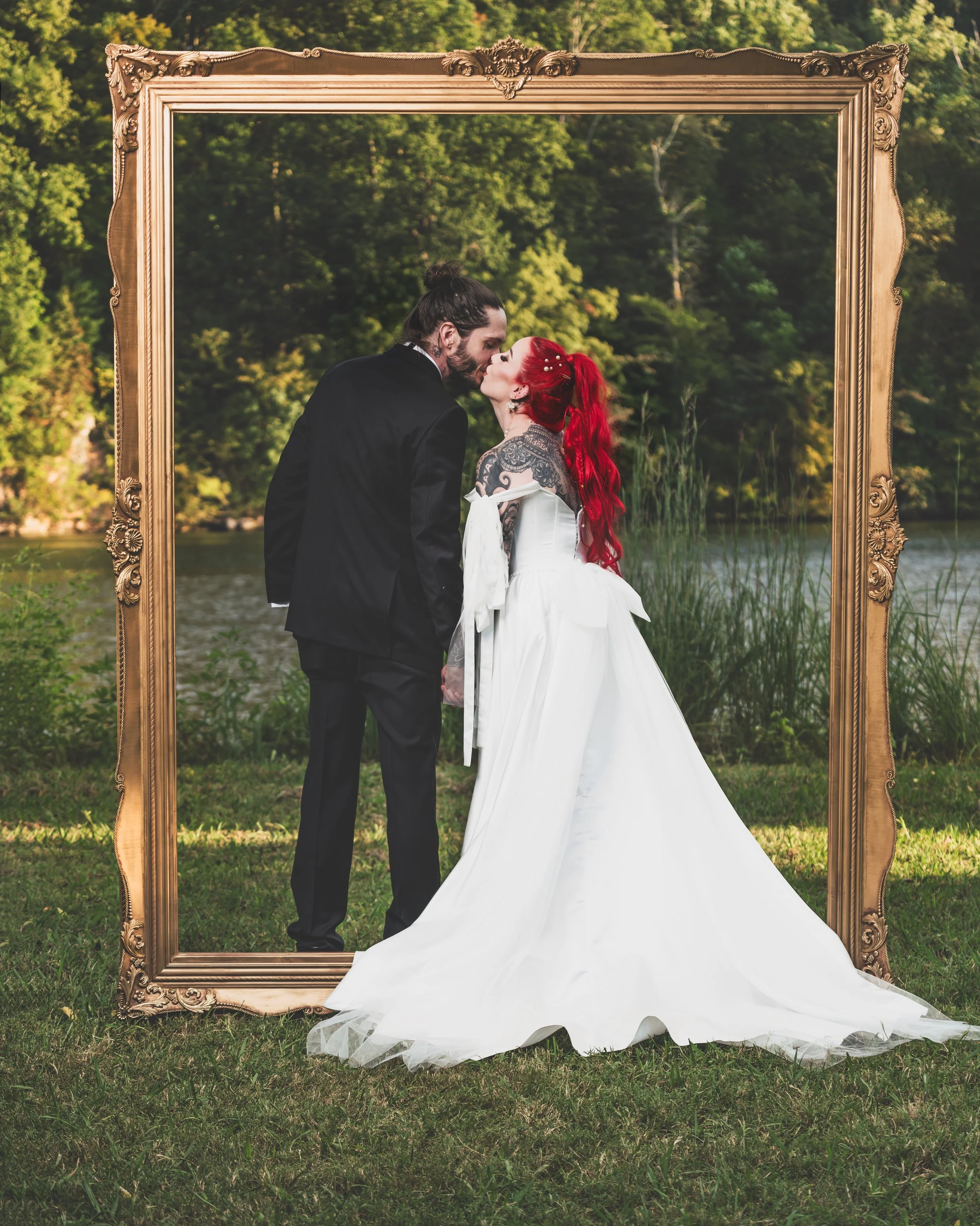 A bride and groom kissing outdoors in a forest by a lake, framed by a large ornate golden picture frame, with the bride in a white gown and vibrant red hair, and the groom in a black suit.