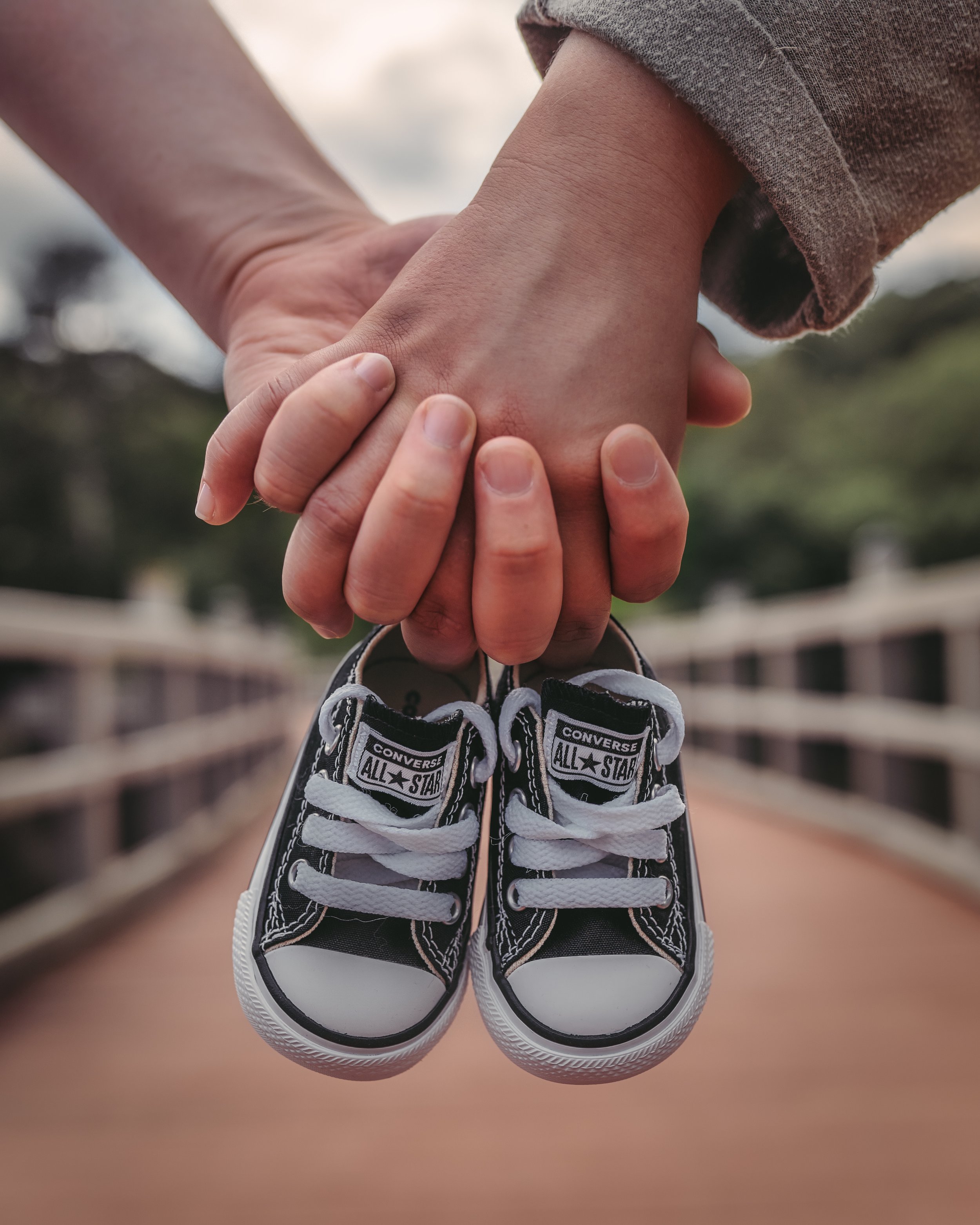 Two hands holding a pair of small black and white Converse sneakers on a blurry outdoor background.