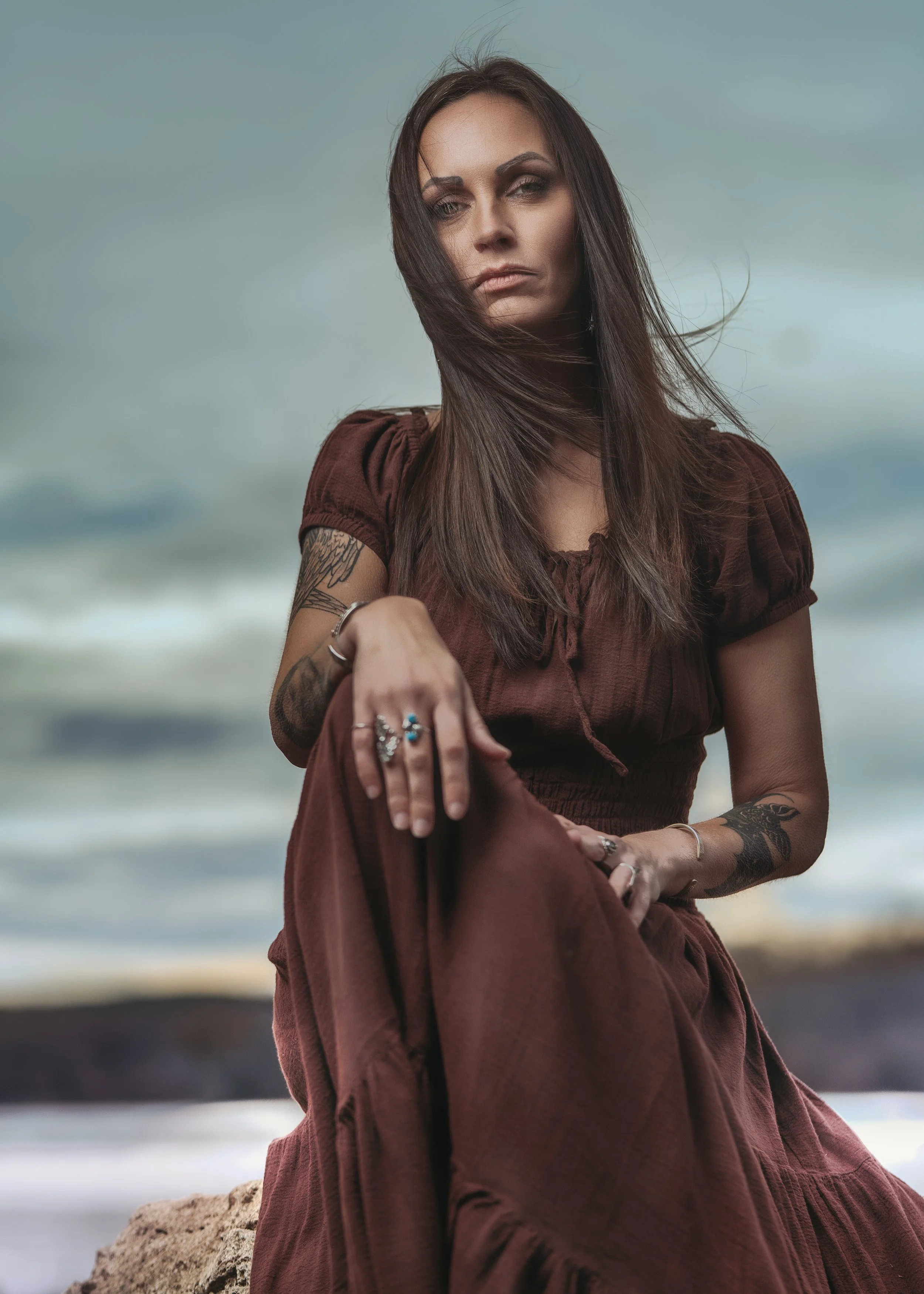 A woman with long dark hair, tattoos on her arms, and multiple rings on her fingers, wearing a brown dress, sitting outdoors against a cloudy sky background.