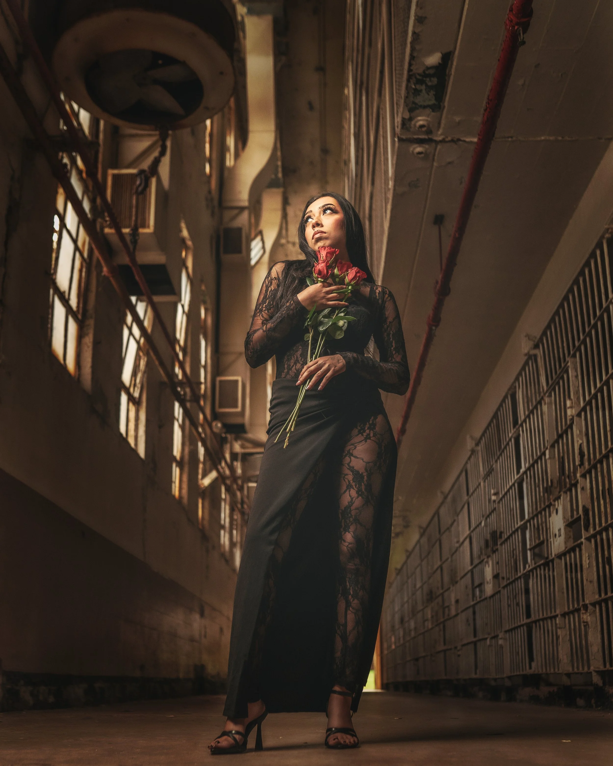 A woman wearing a black lace dress with a high slit, holding a bouquet of pink roses, standing in an industrial corridor with rusted pipes and windows with broken glass.