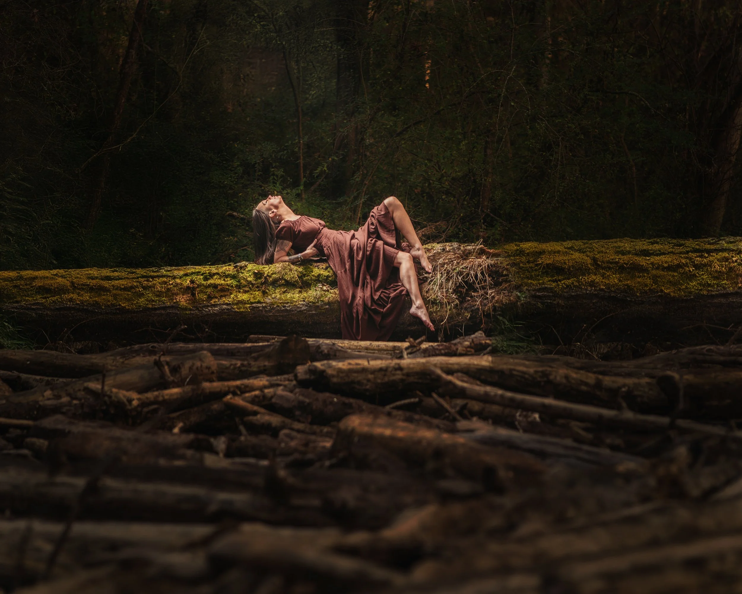A woman in a long brown dress lying on a moss-covered log in a dense forest, with her head tilted back and eyes closed.