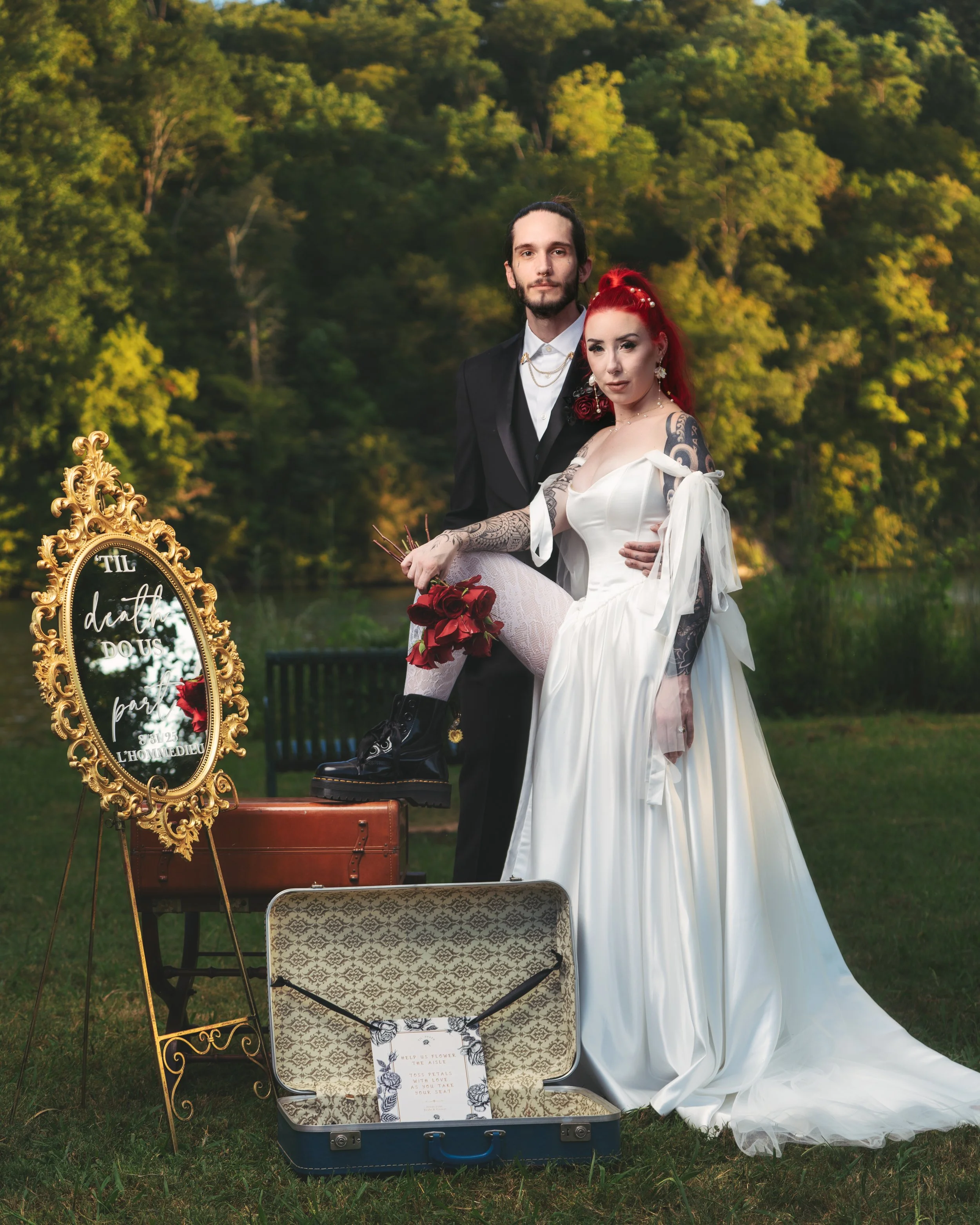 A newlywed couple standing outdoors near a river and trees during sunset, with vintage accessories and a mirror with a wedding message.