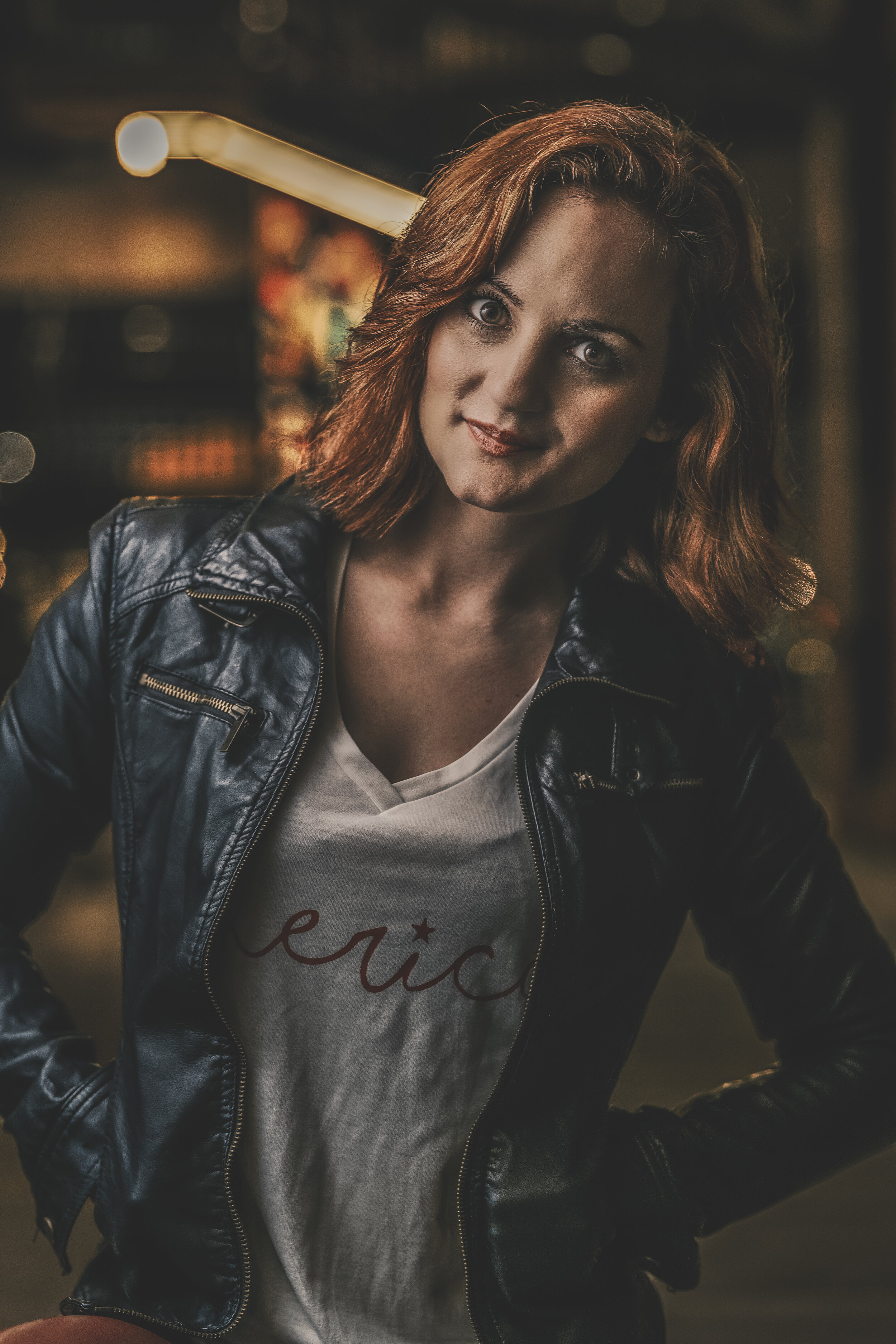 Woman with red hair in a black leather jacket and a white shirt with text, smiling at the camera in an urban setting.