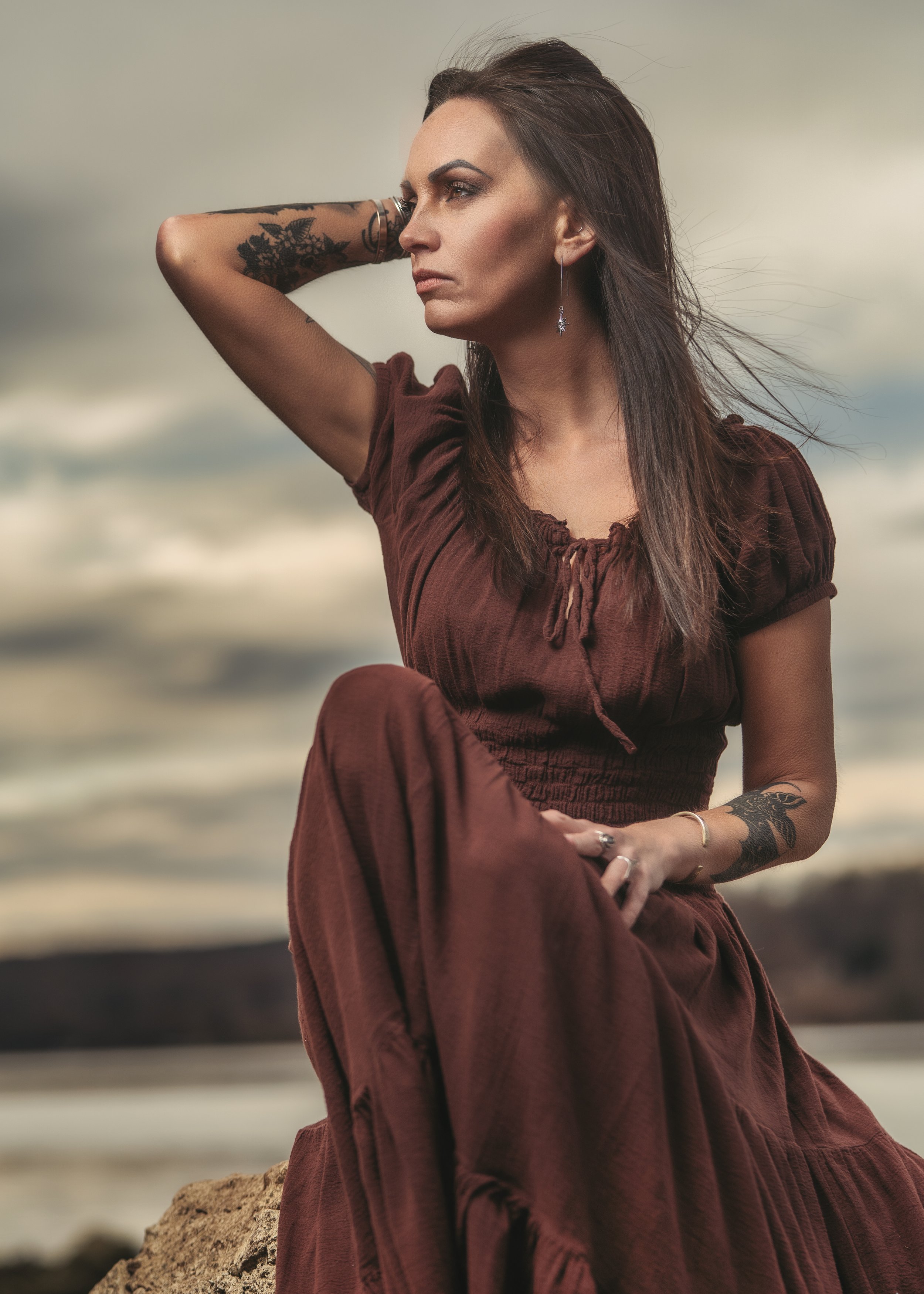 A woman dressed in a long, dark reddish-brown dress sitting outdoors on a rock near the water, with cloudy sky backgrounds. She has long dark hair, tattoos on her arms, and is looking to the side with her hand resting behind her head.