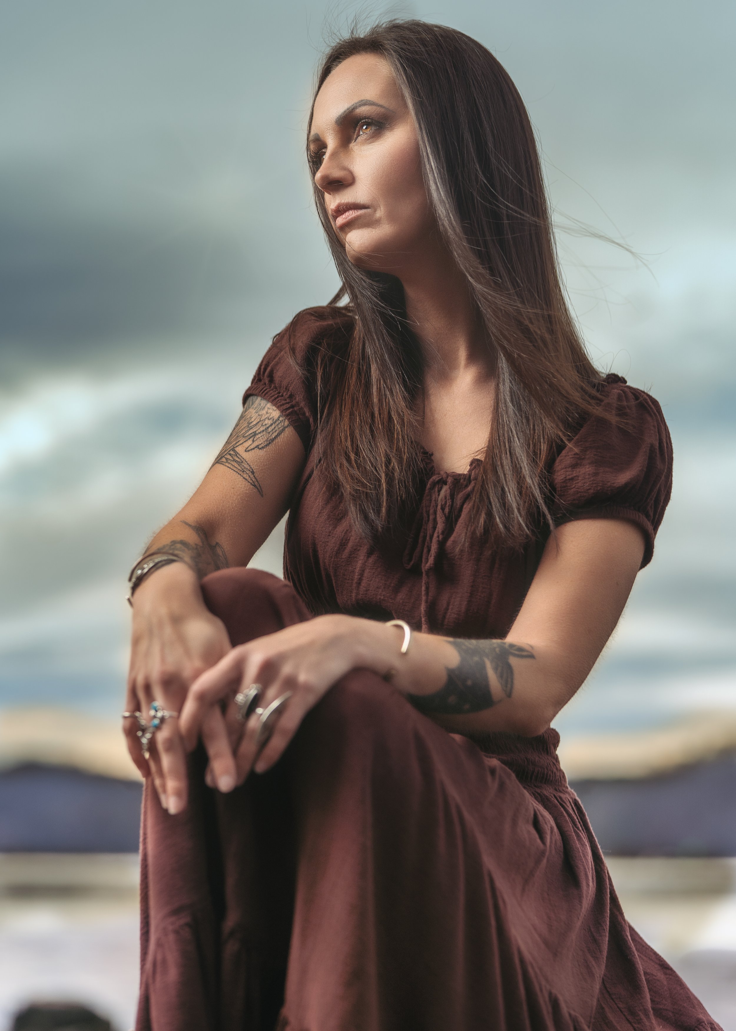 A woman with long brown hair, wearing a brown short-sleeved dress, sitting outdoors with a cloudy sky background, looking to the side.