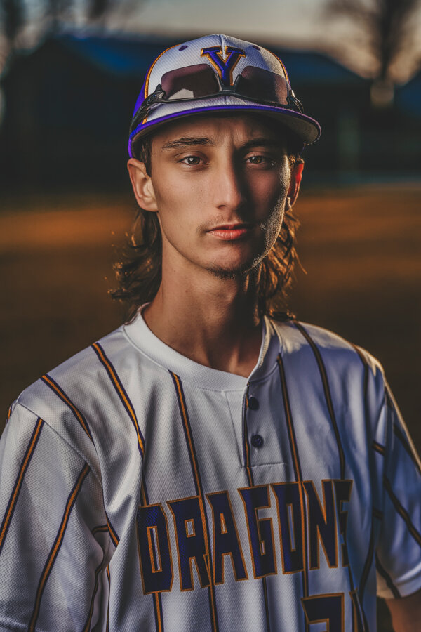 A baseball player in a white and purple jersey with "Dragons" written on it, wearing a baseball cap and sunglasses resting on the cap. The background is blurred, indicating an outdoor setting.