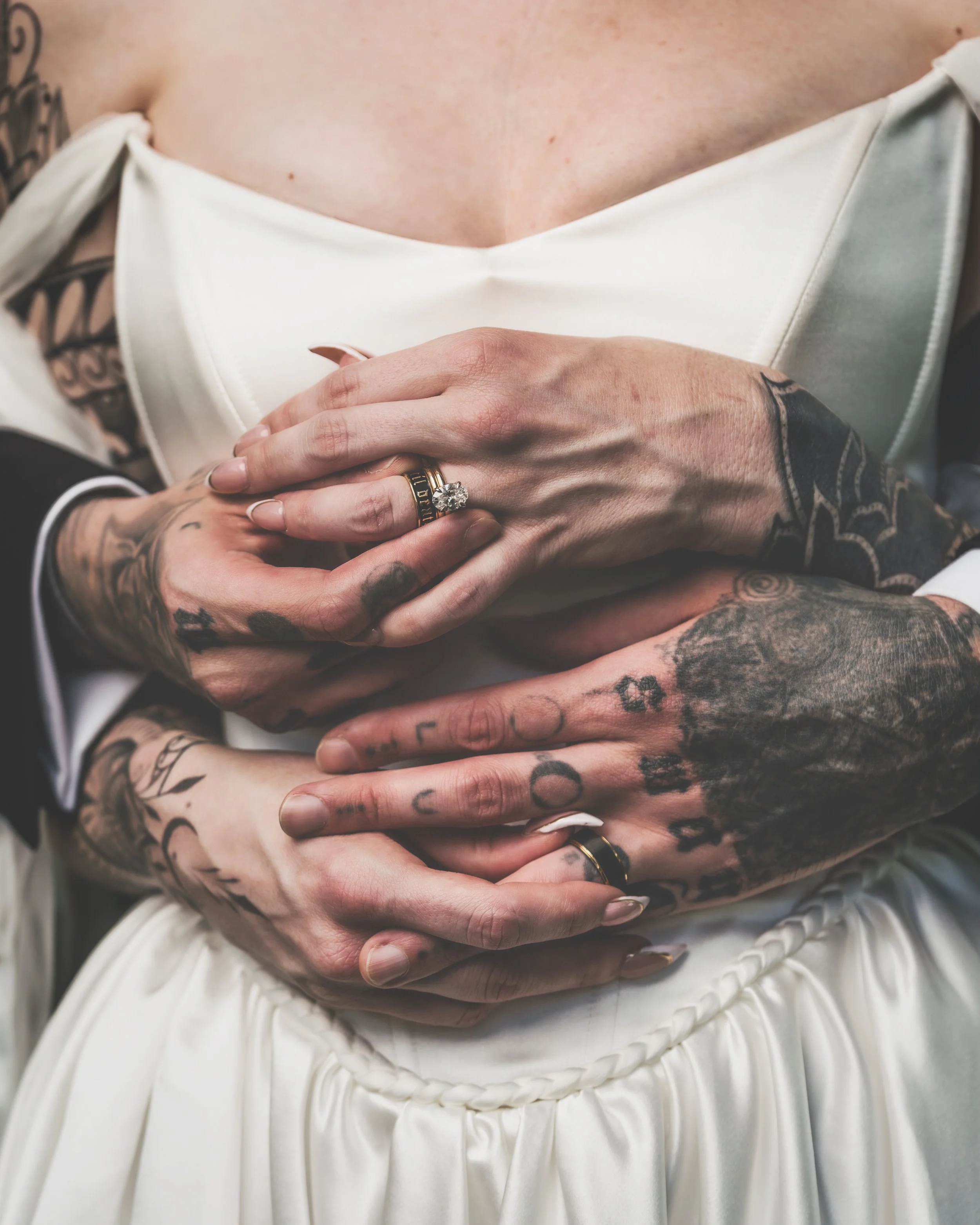 Close-up of a couple's hands with tattoos, holding each other over a white dress, showcasing wedding rings.