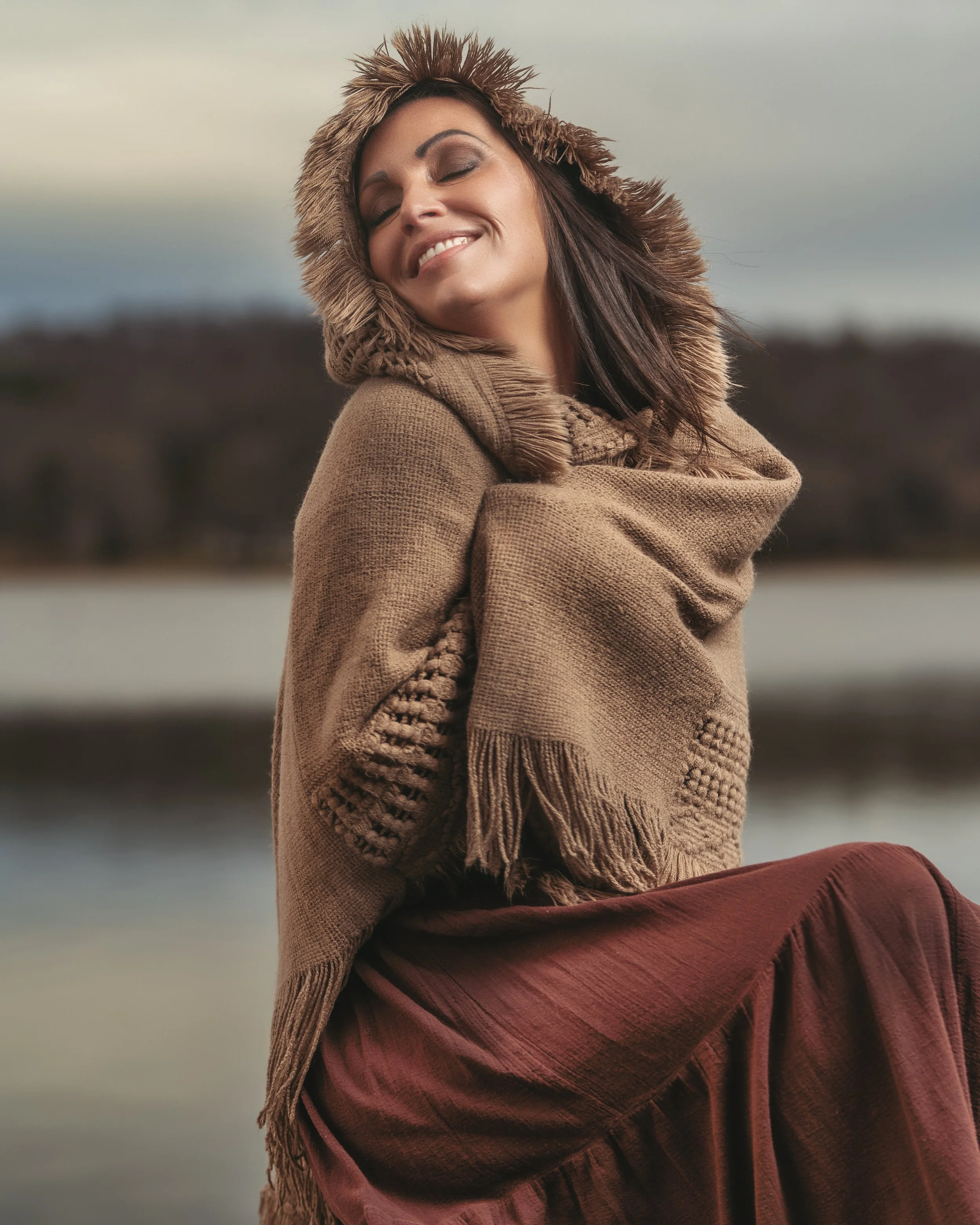 A woman smiling with eyes closed, wearing a brown fur-lined hooded coat and a large beige scarf, sitting outdoors near a body of water with a blurred natural landscape in the background.