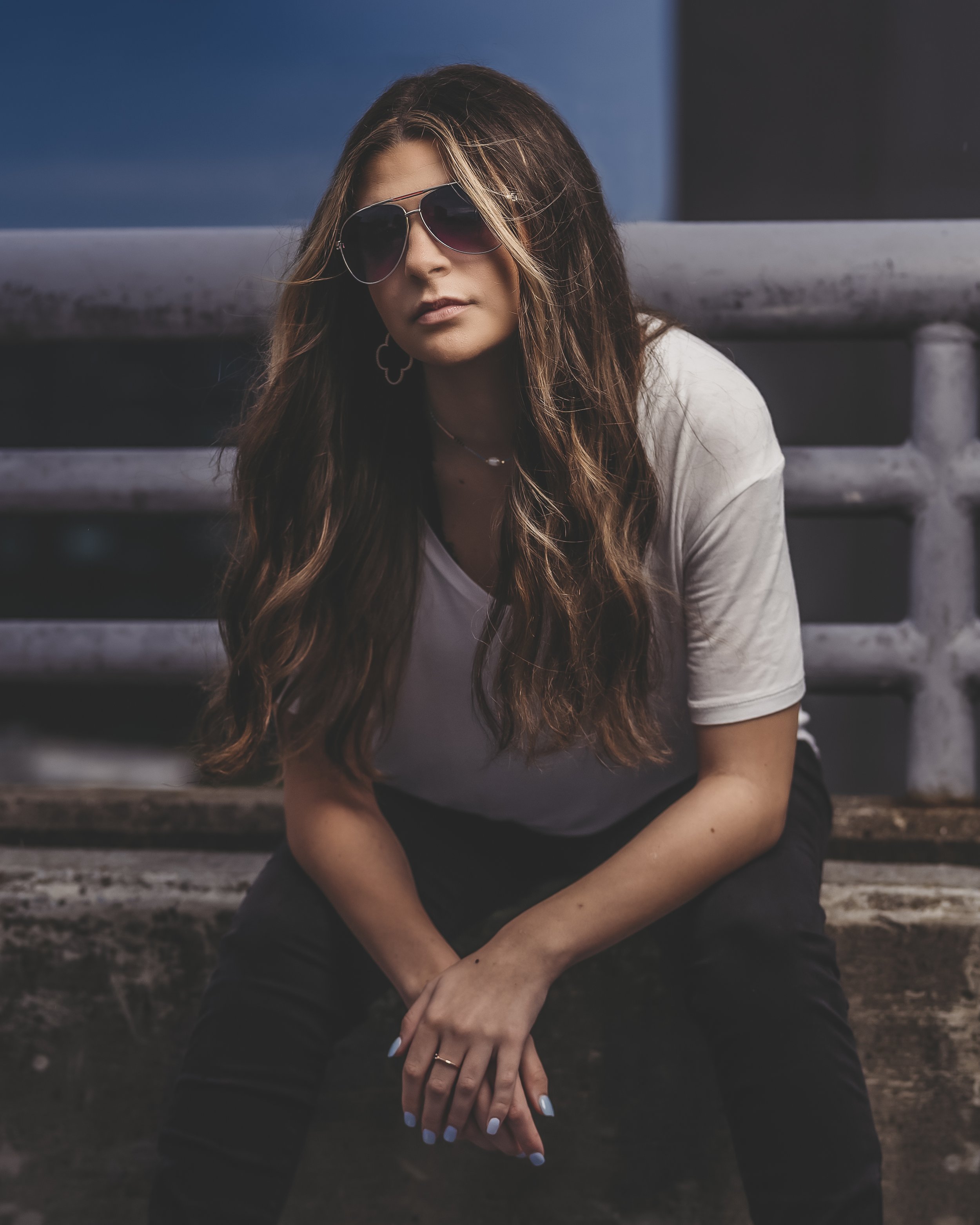 Woman with long hair wearing sunglasses, a white shirt, and black pants sitting on a concrete ledge.