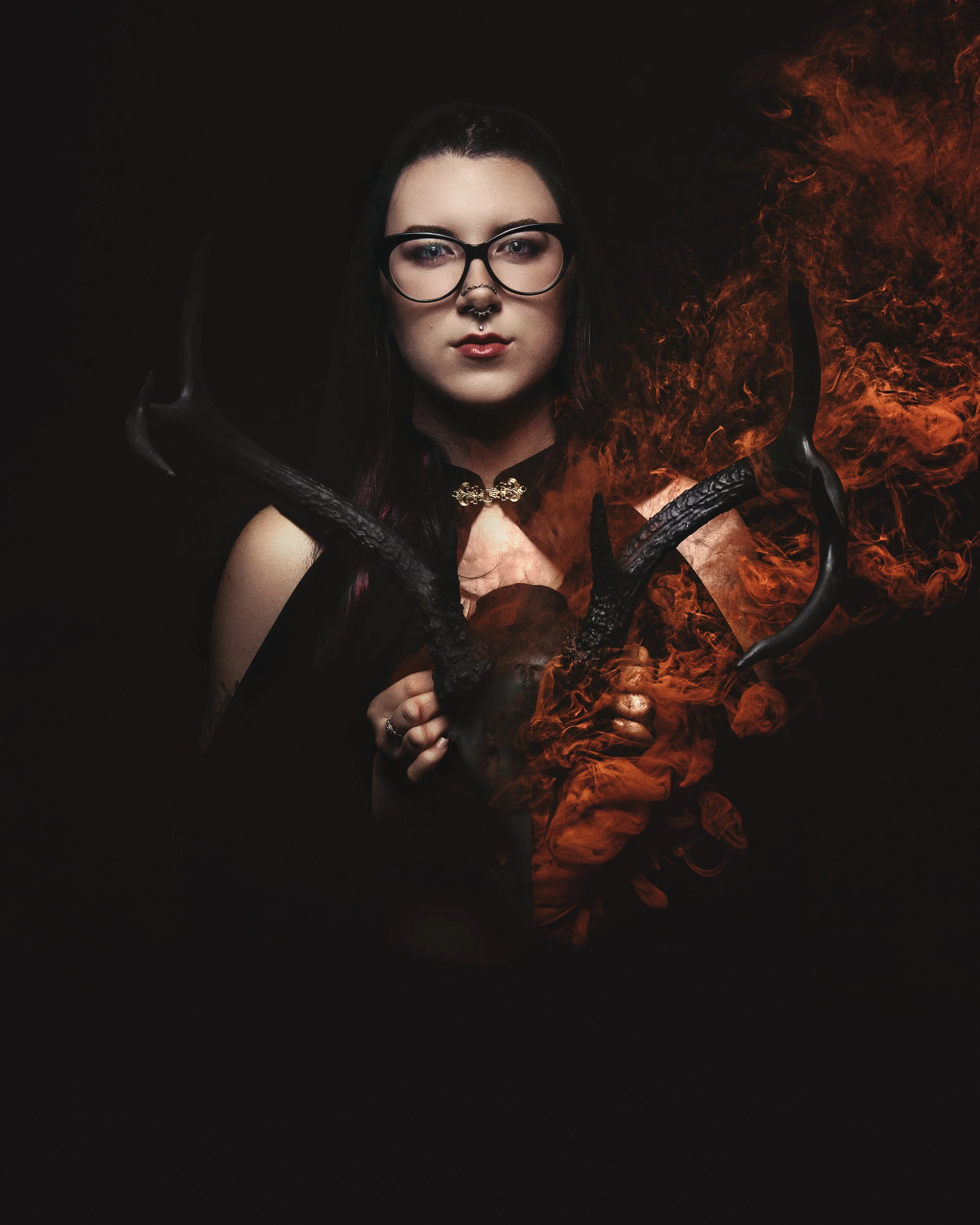 A woman with glasses and dark hair holding a skull with black antlers, with flames emanating from the skull, against a dark background.