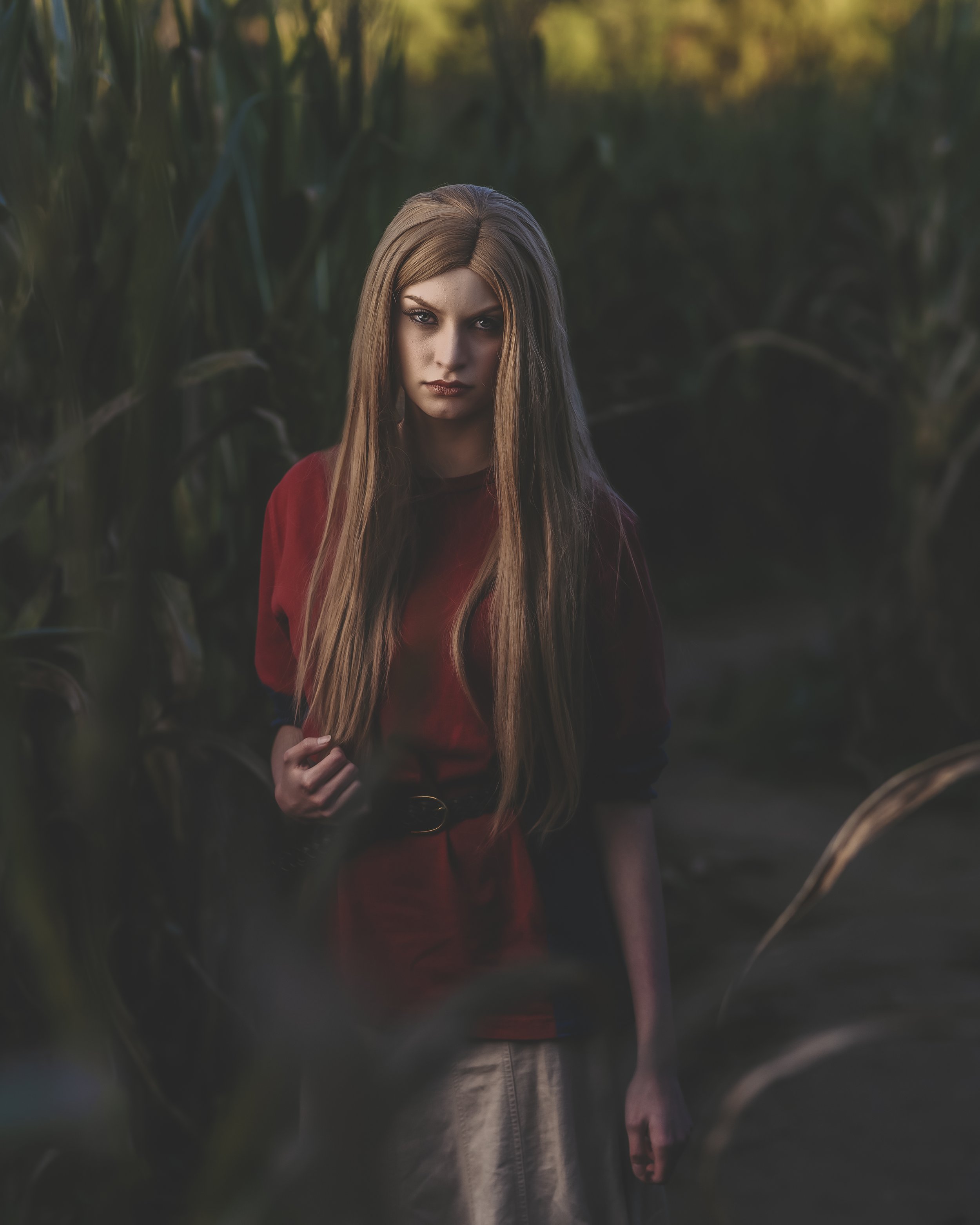 Person with long hair wearing a red shirt standing in a cornfield, with low lighting and natural surroundings.