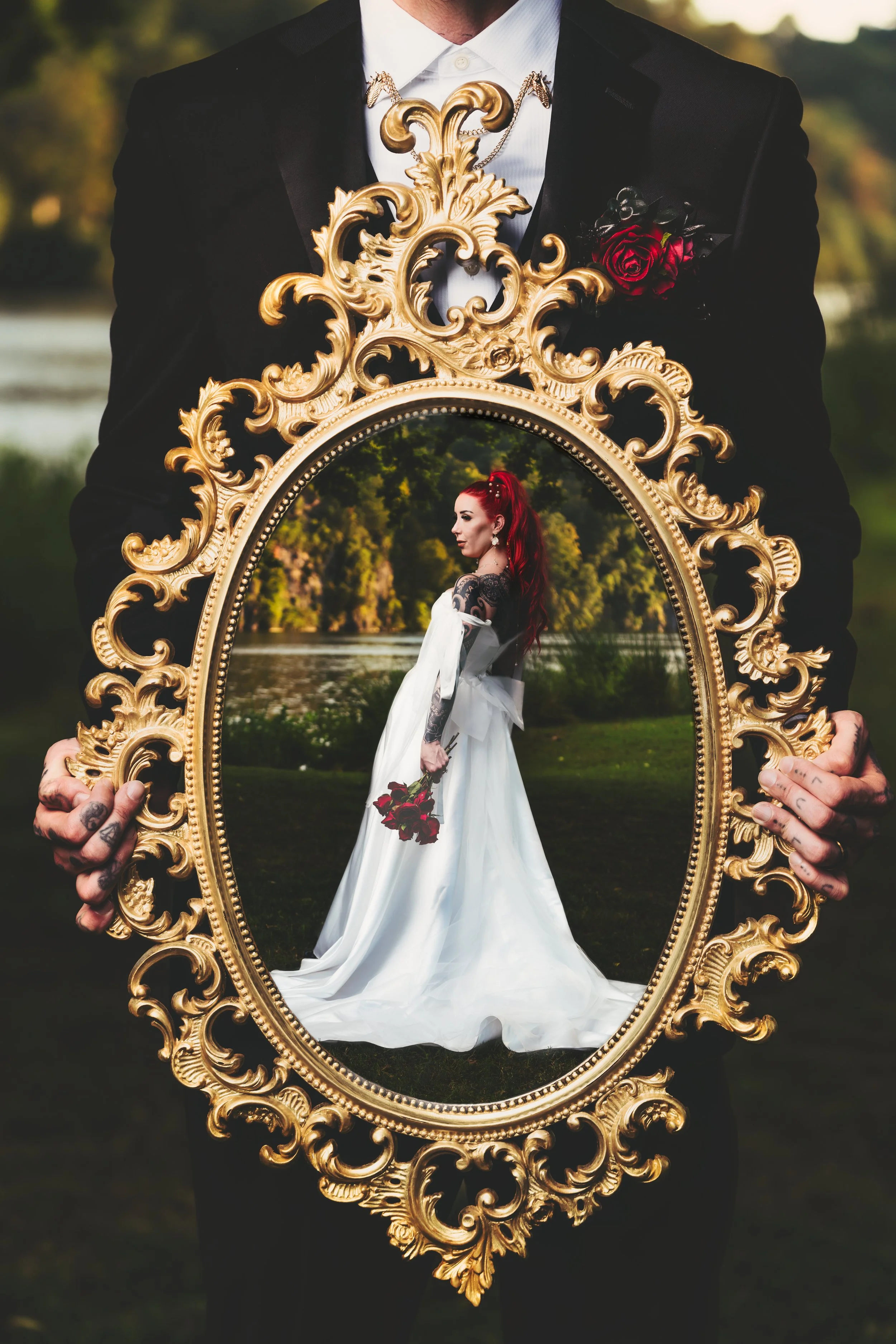 A person in a suit holding an ornate gold frame that reflects a woman in a wedding dress standing outdoors by a river with lush green trees in the background.