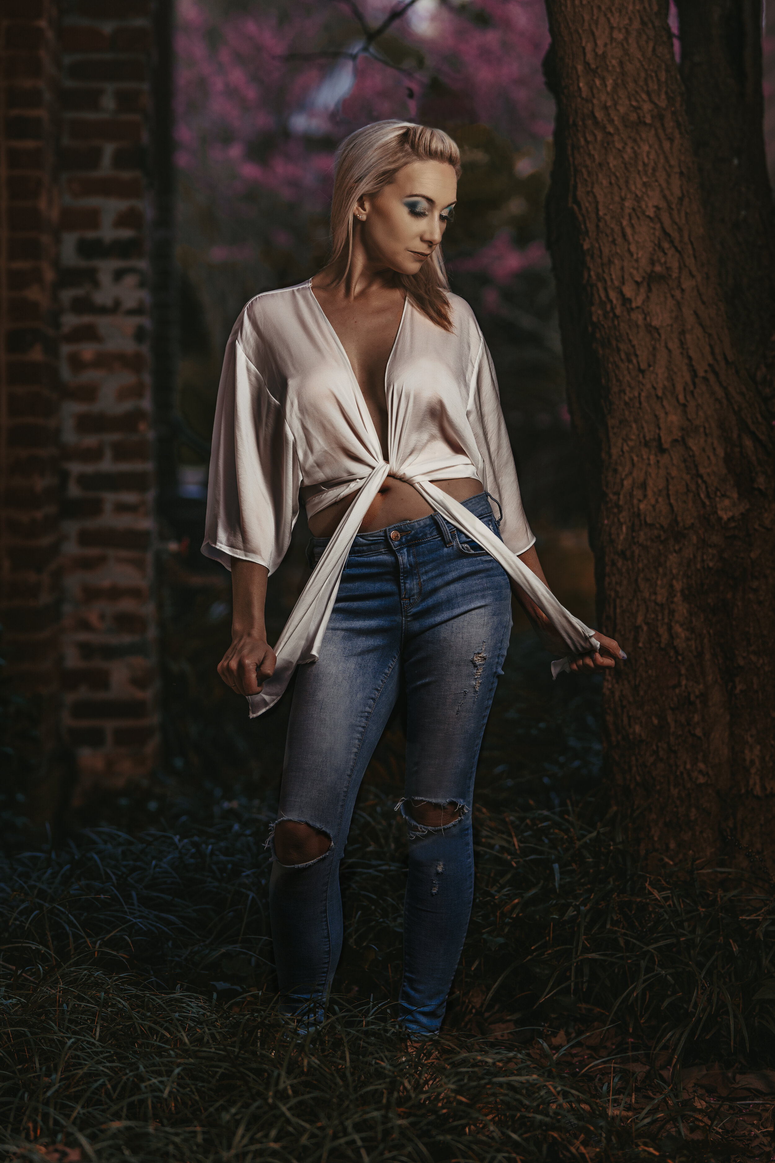 Woman in ripped jeans and white blouse standing outdoors near tree, with blurred pink flowers in the background.