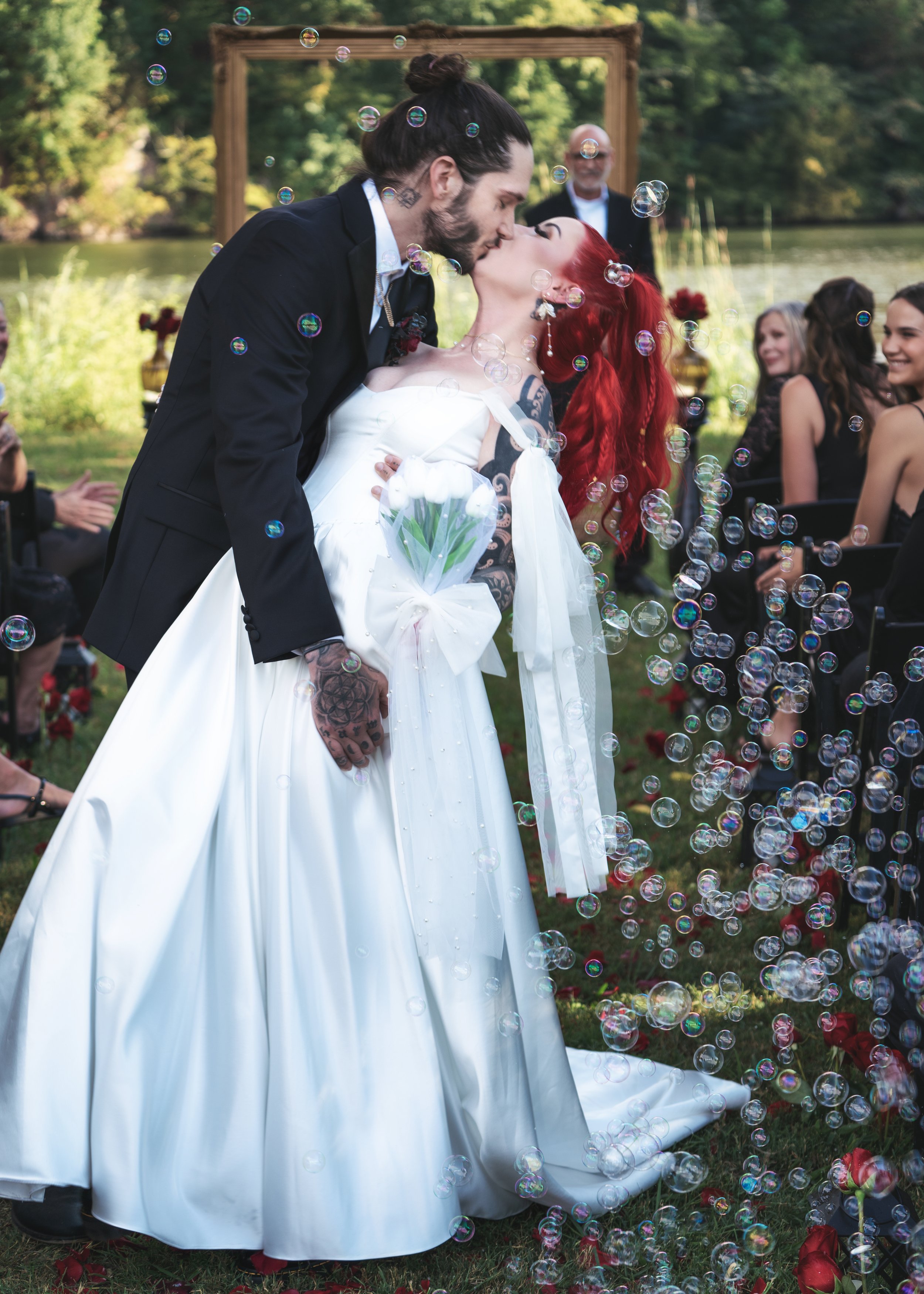 Couple kissing at their outdoor wedding ceremony, with bubbles floating around and guests seated nearby.