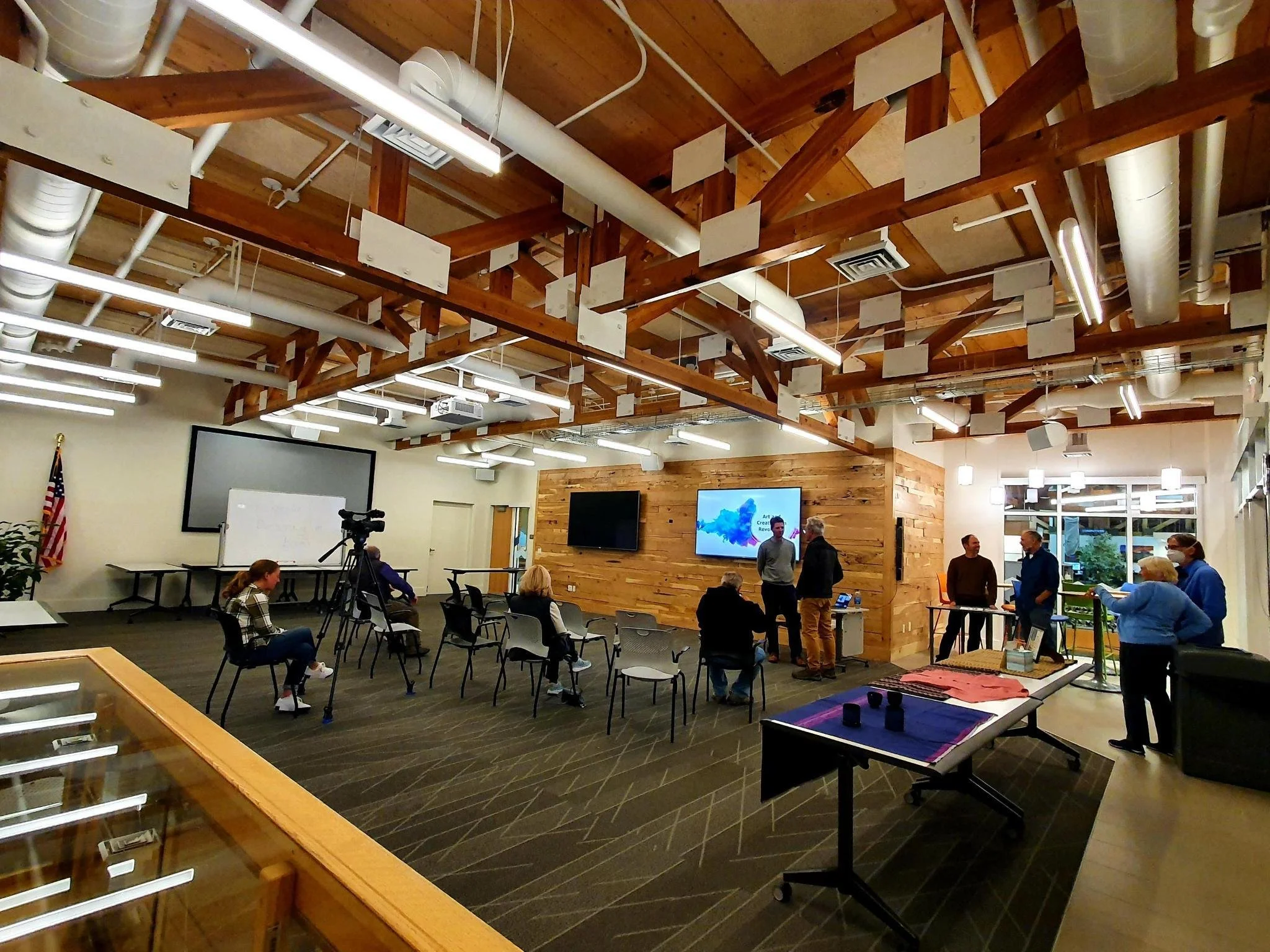 Interior of a modern meeting room with wooden beams, people gathered, chairs, American flag, cameras, and screens.
