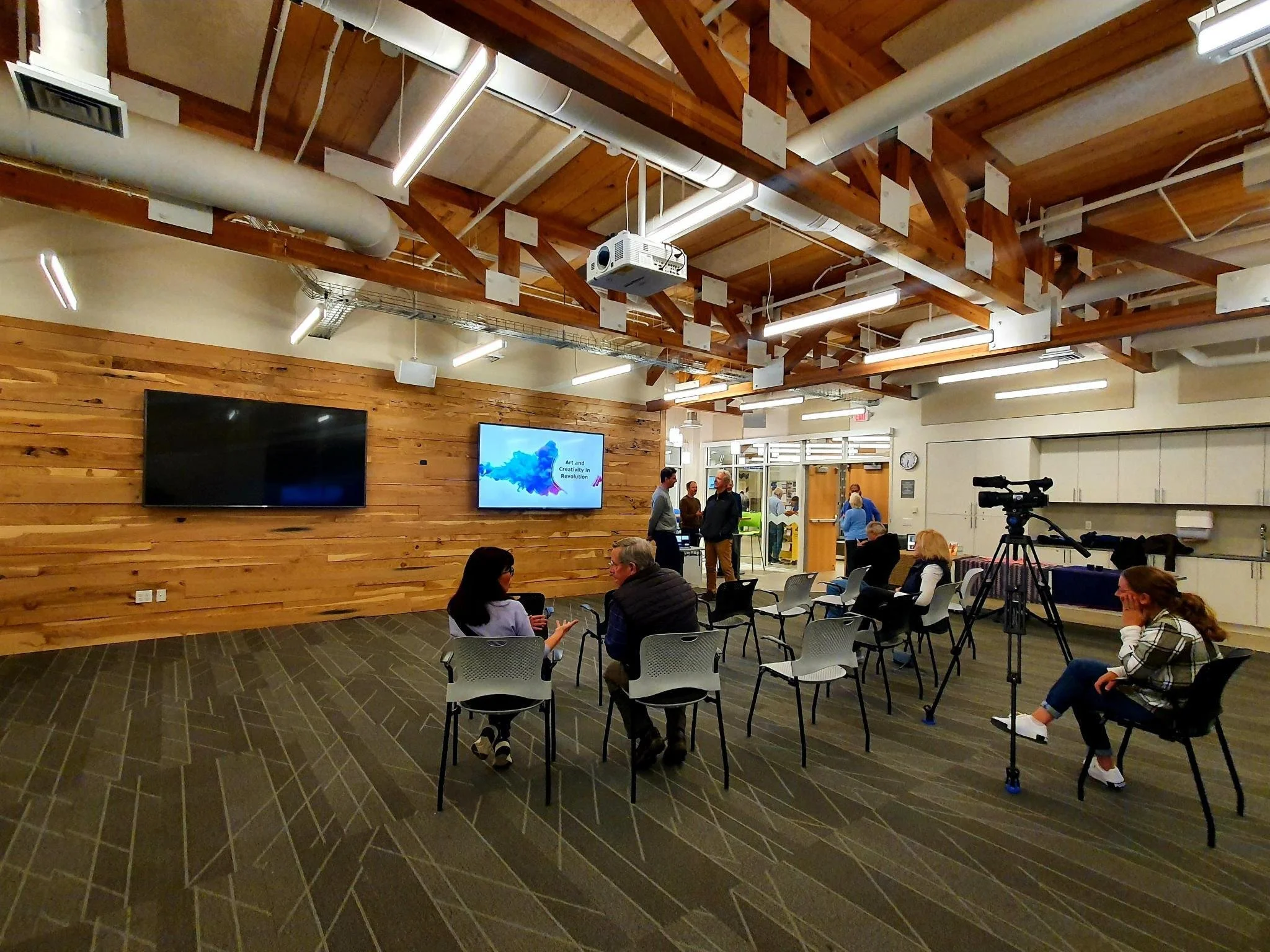 Conference room with people, chairs, camera, screens, and exposed wooden beams
