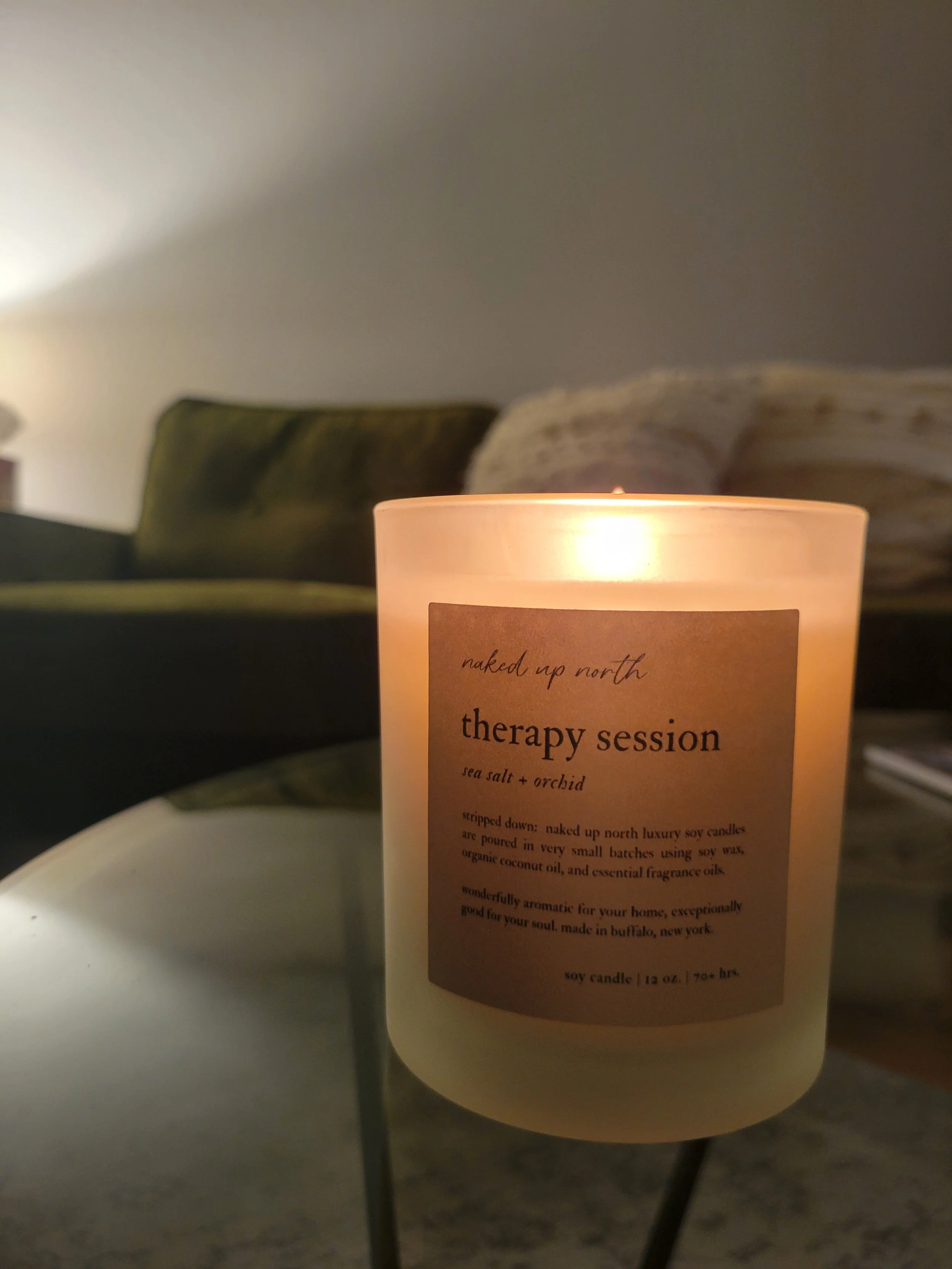 A lit candle on a table with a label reading 'naked up north therapy session sea salt + orchid' with a blurred background of a couch and wall.