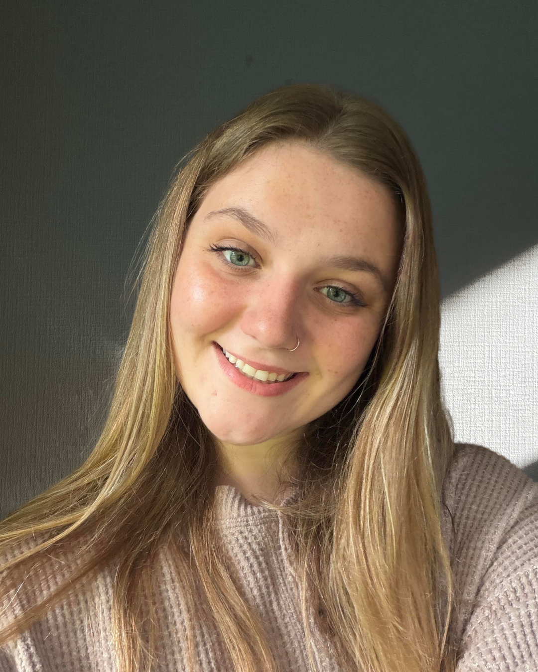 Close-up of a young woman's face smiling, with long blonde hair, wearing a pink sweater, in a room with a wooden headboard in the background.