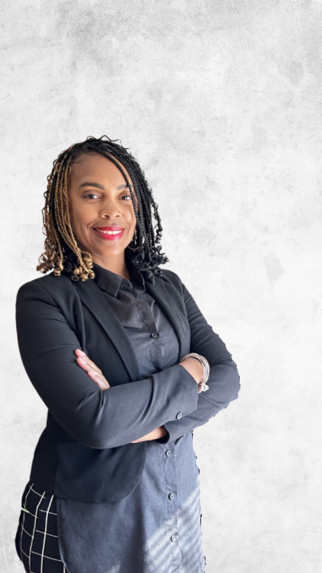 A woman with braided hair standing in an office with her arms crossed, smiling, dressed in a black blazer and checkered pants, with framed diplomas on the wall, a potted plant on the desk, and an office chair behind her.