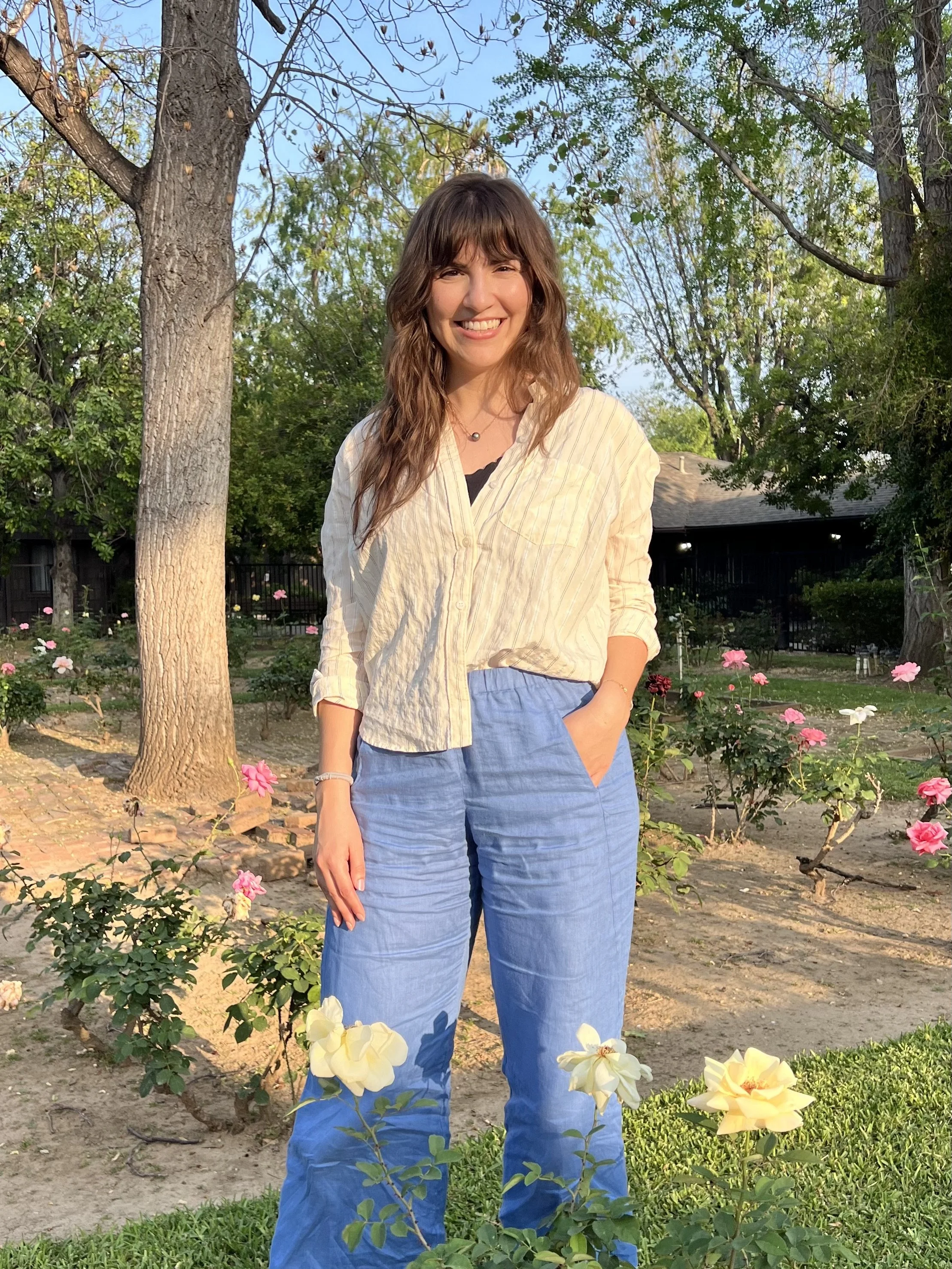 A young woman with long wavy brown hair, smiling, wearing a cream-colored shirt and blue pants, standing outdoors in a garden with flowers and trees, during late afternoon sunlight.