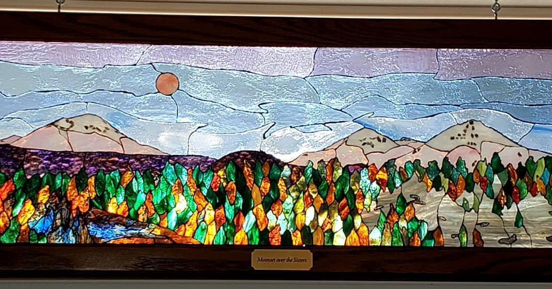 This horizontally made glass mosaic is a rendition  of the view from Hwy 20 on the way to Bend, OR.  It's of the 3 Sisters at dawn with the setting of a full moon.  The early morning pink sky casts its light on the mountains as lakes and high desert 