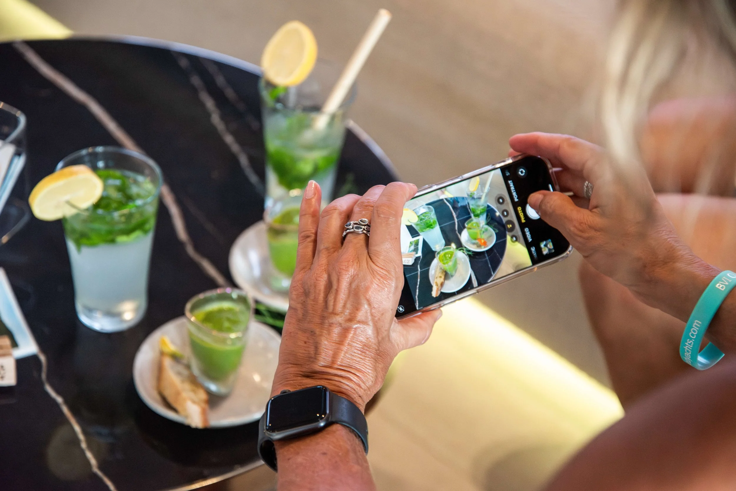 A woman taking a photo of drinks with a smartphone. The drinks are mint and lemon mojitos on a black marble table.