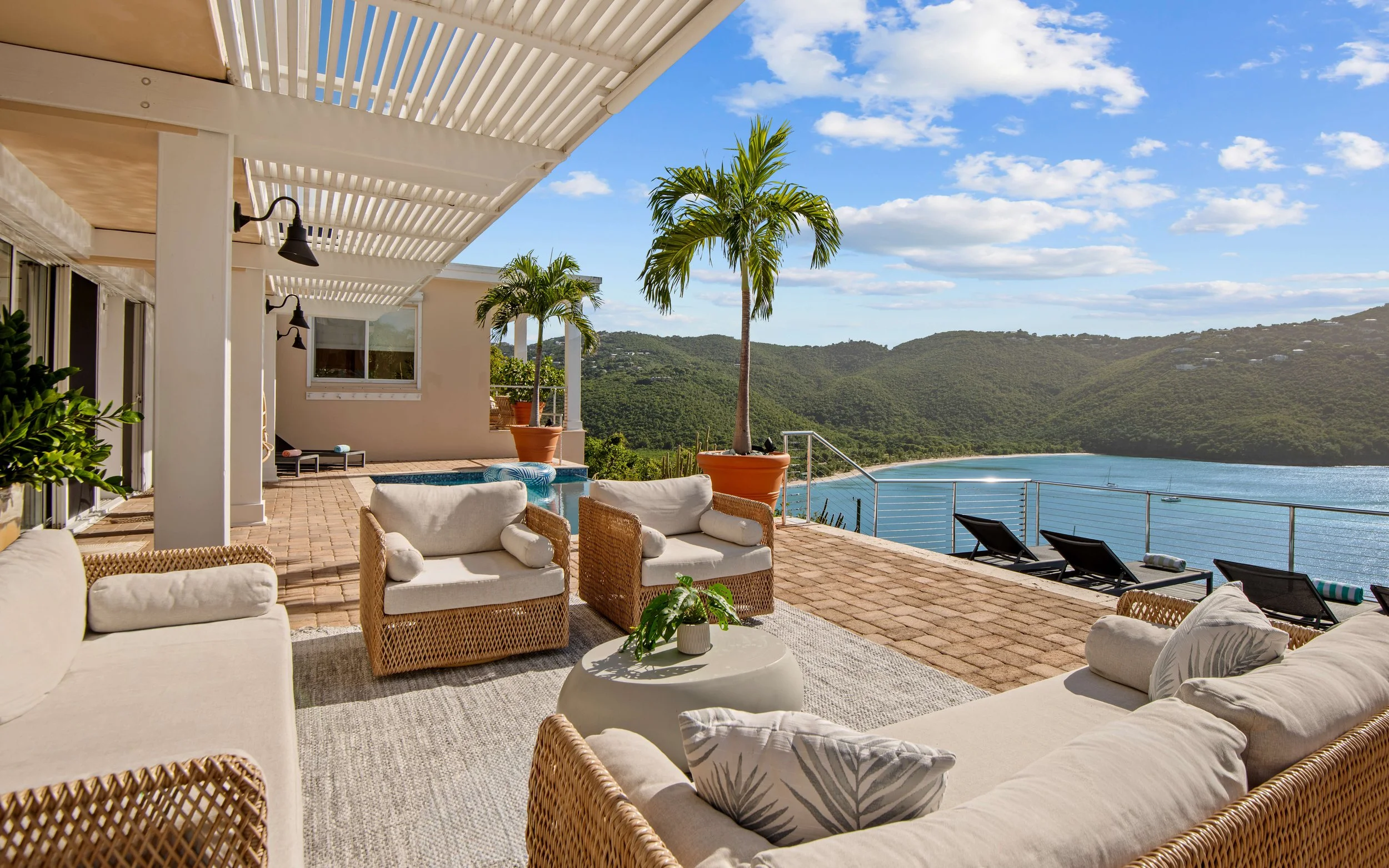 An outdoor patio with rattan and cushioned seating overlooking a bay with a small sailboat, mountains in the distance, and a partly cloudy sky.