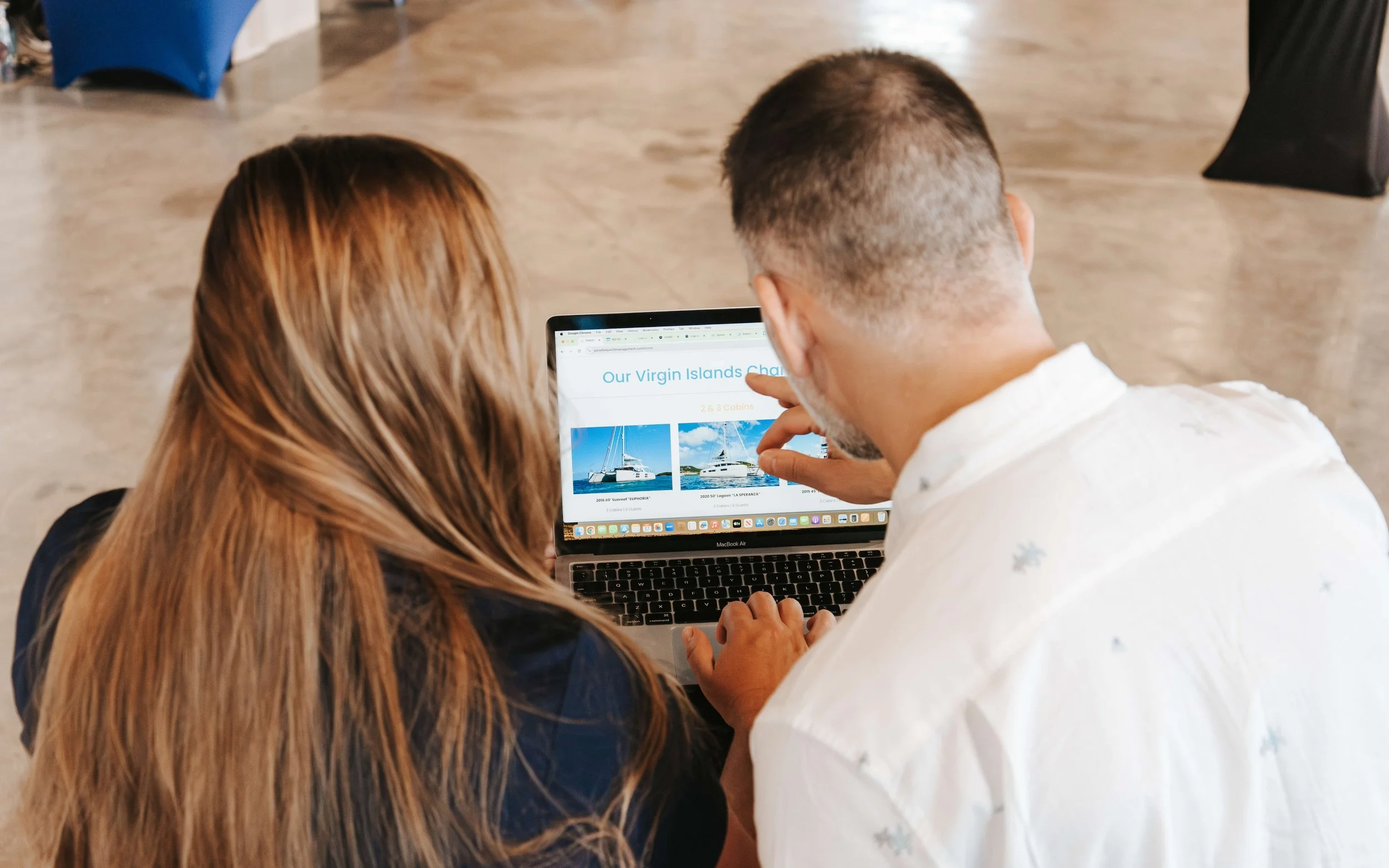 Two people viewing a laptop screen with pictures of sailboats for sale or rental.