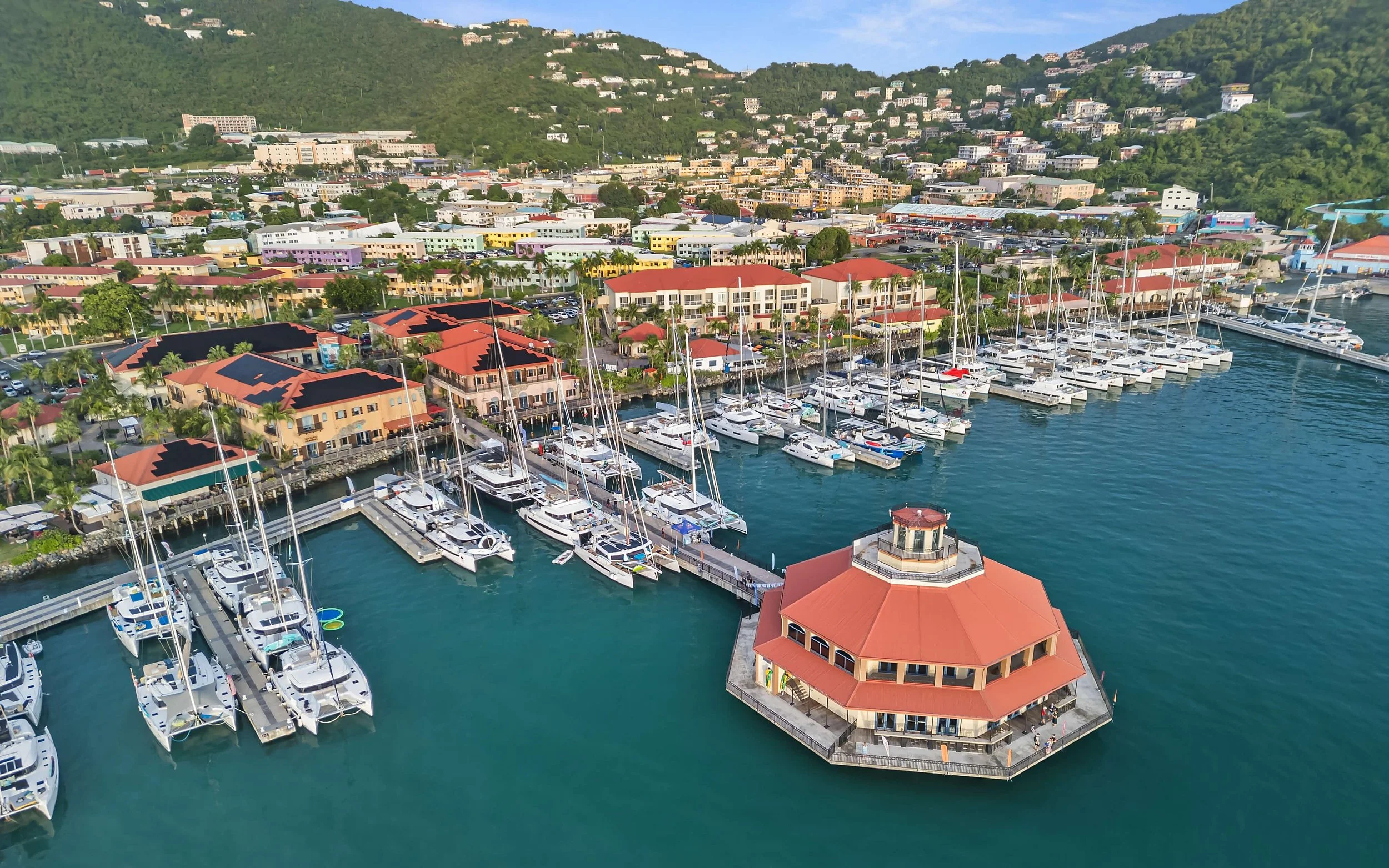 Aerial view of a marina with numerous sailboats docked, a waterfront building with a red roof, colorful buildings and houses on a hillside in the background.