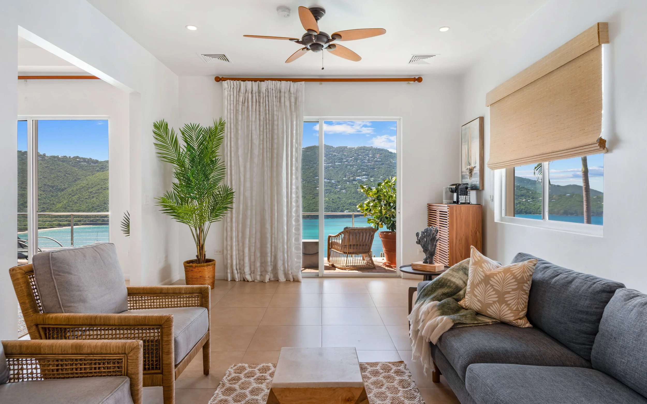 Living room with sliding glass door leading to balcony overlooking a water body and green hills, with tropical plants and couches.