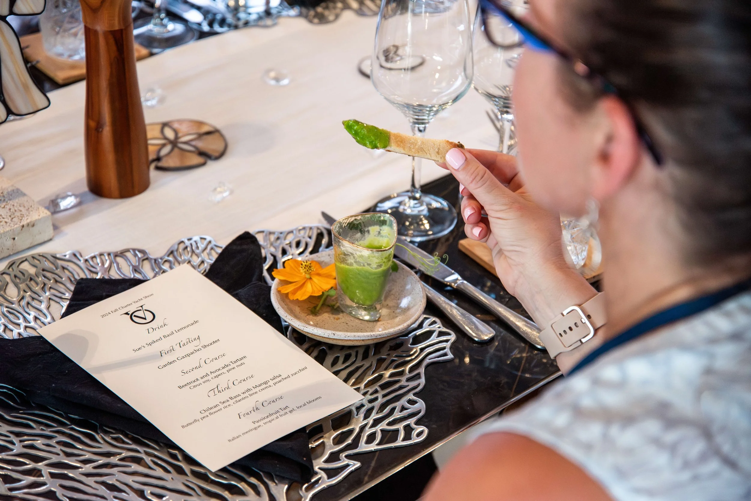 A woman at a dining table holding a toast with green spread, with an appetizer glass and a menu on the table.