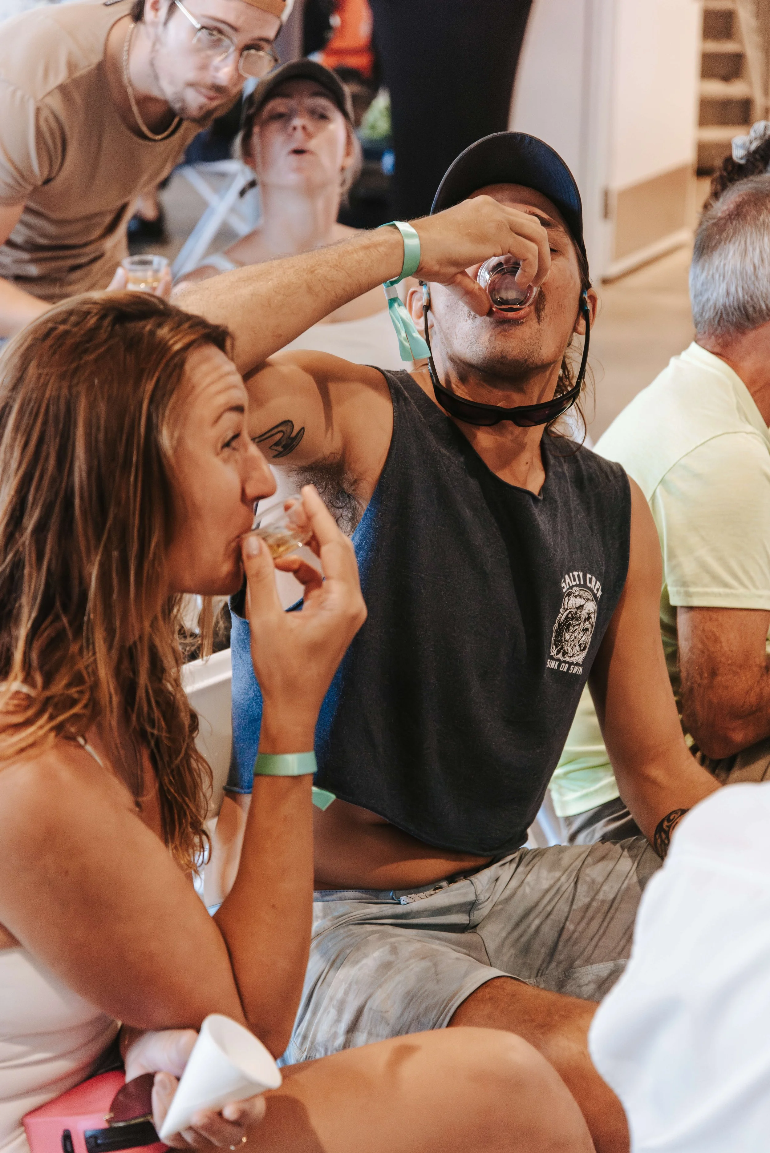 Group of people at a social gathering, with some drinking, one person pouring a drink into his mouth, others watching and socializing