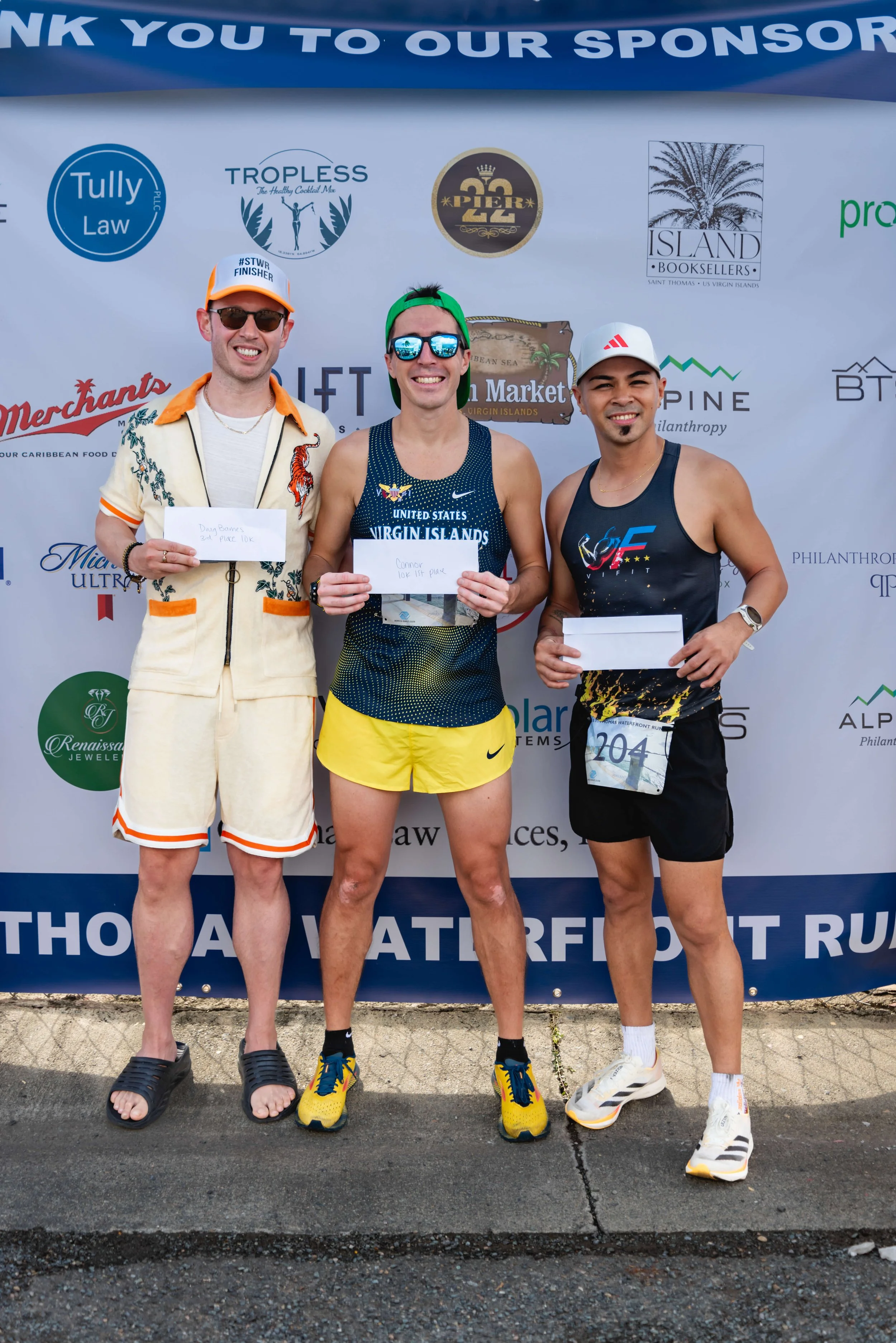 Three male runners standing in front of a sponsorship banner at a race event, holding envelopes with handwritten messages, smiling after completing a marathon or race.