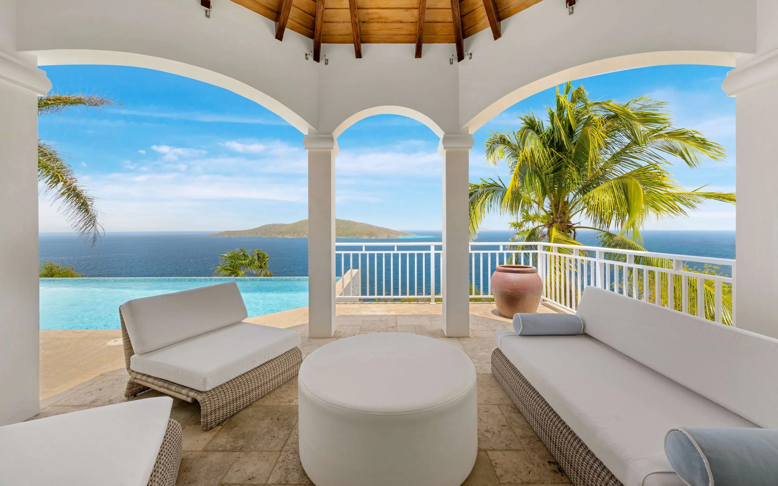 A covered patio with white wicker couches, a round table, and large pots, overlooking the ocean with a view of a small island and palm trees.