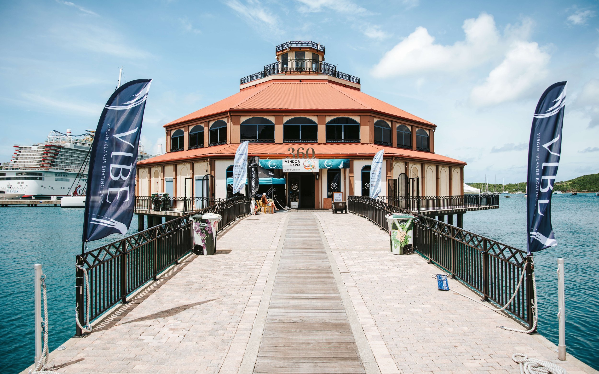 A circular building with a red roof and large windows situated over water, with flags and banners advertising a vendor exposition on a sunny day.