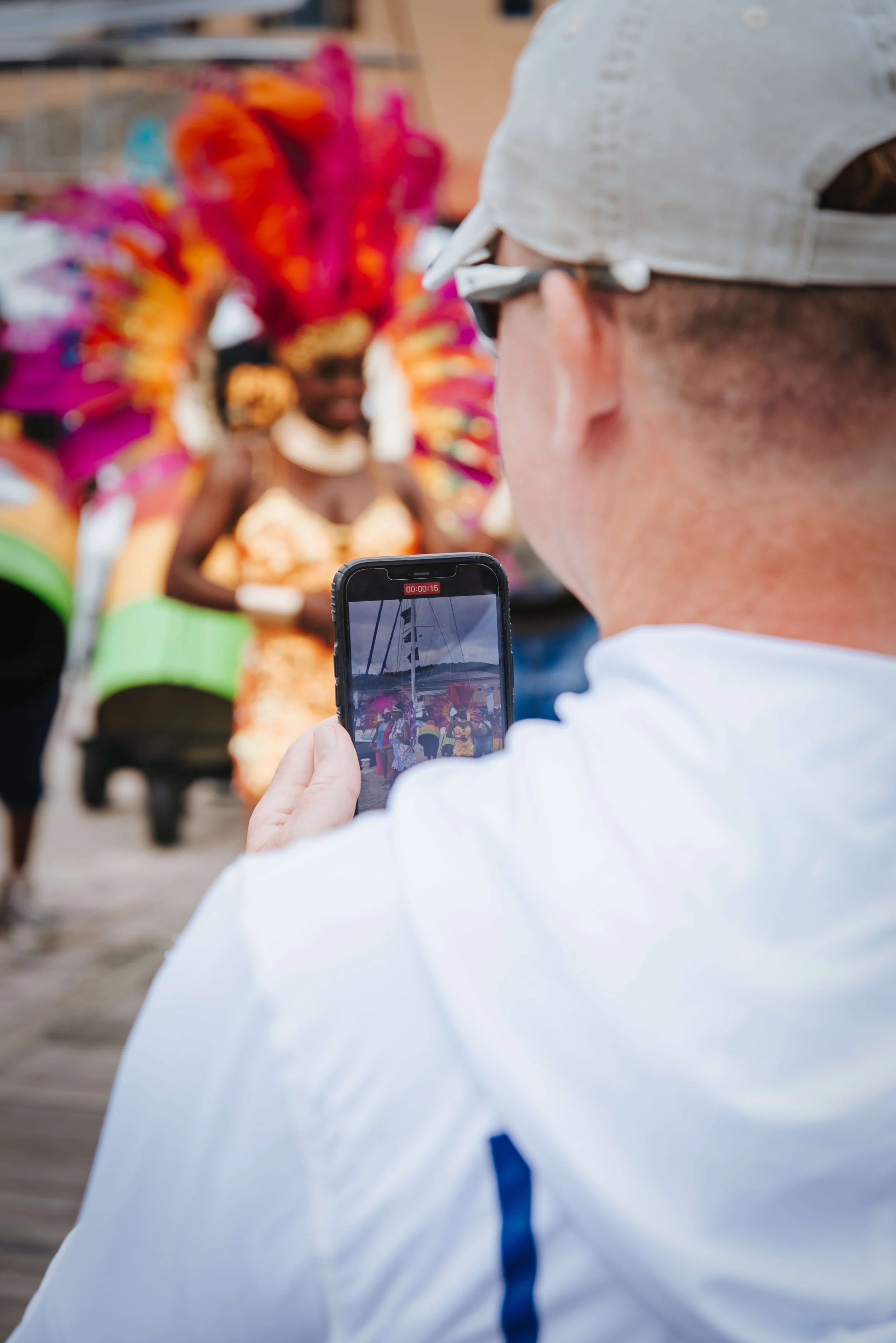 Person filming a performer in colorful costume with large feathered headdress using a smartphone at a street event.