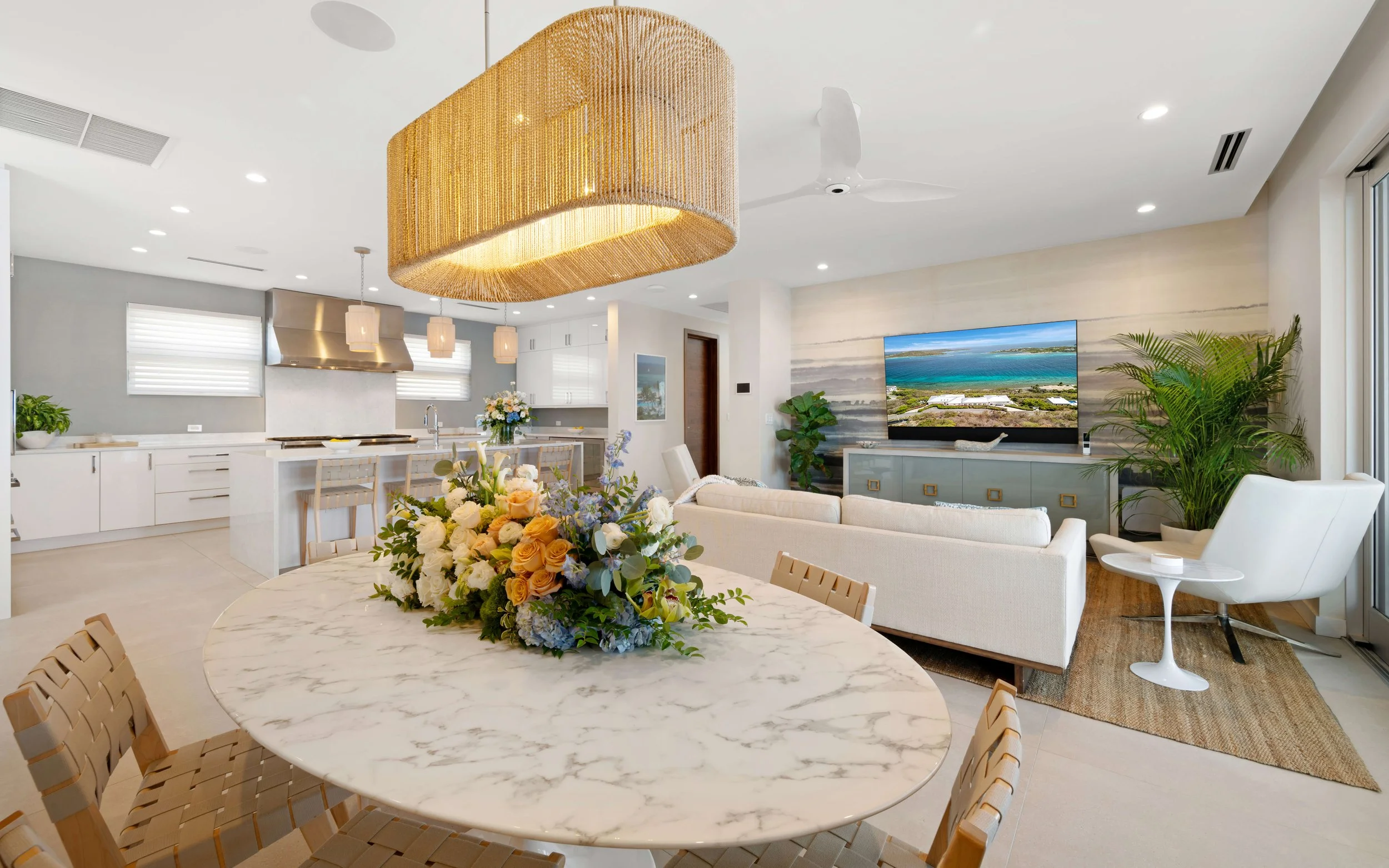 Open-concept living and kitchen area with a white marble dining table, floral centerpiece, white sofa, large flat-screen TV, and lush green plants, with windows and modern decor.