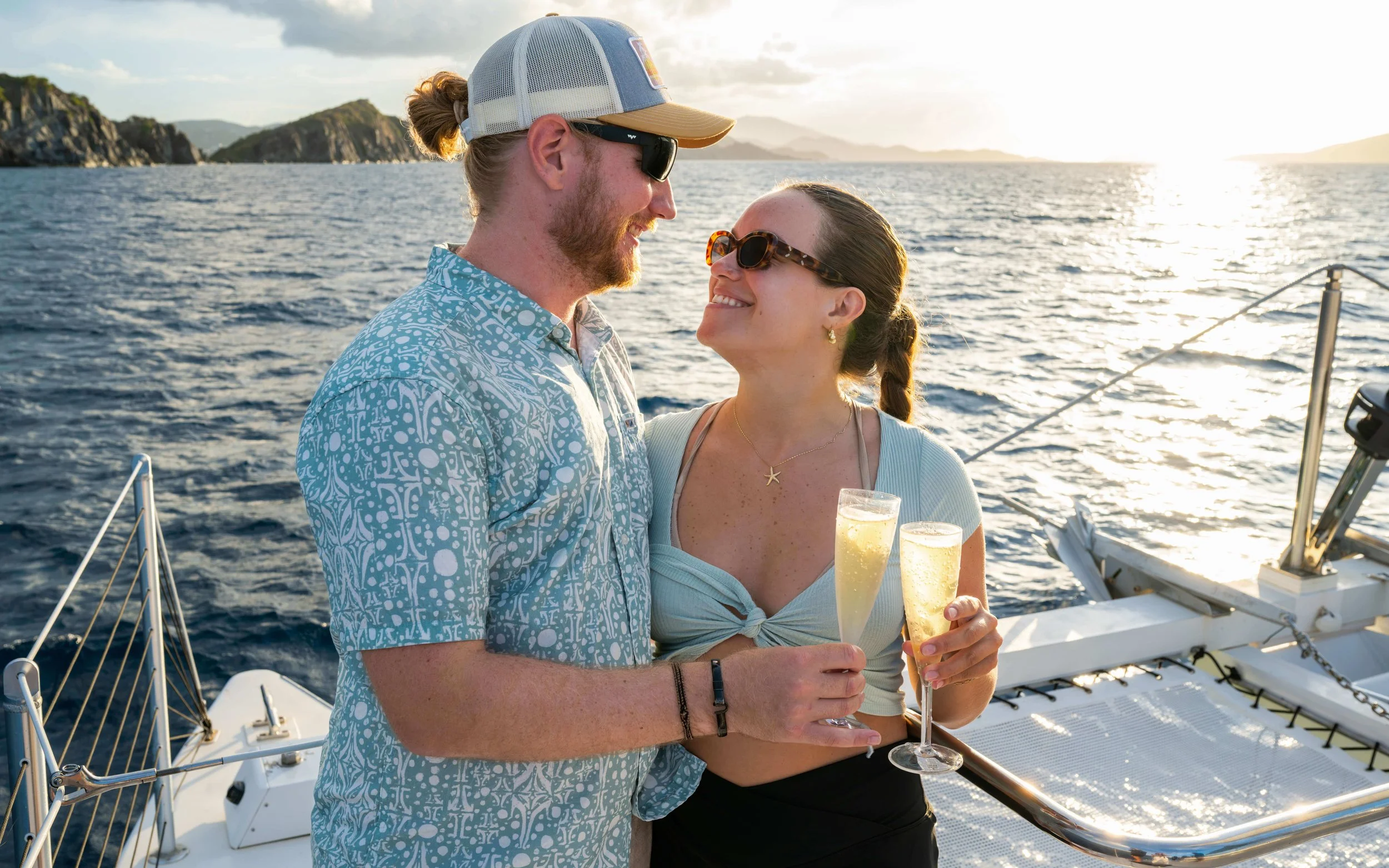 A man and woman on a boat holding glasses of champagne, smiling and looking at each other as the sun sets over the water with islands in the background.