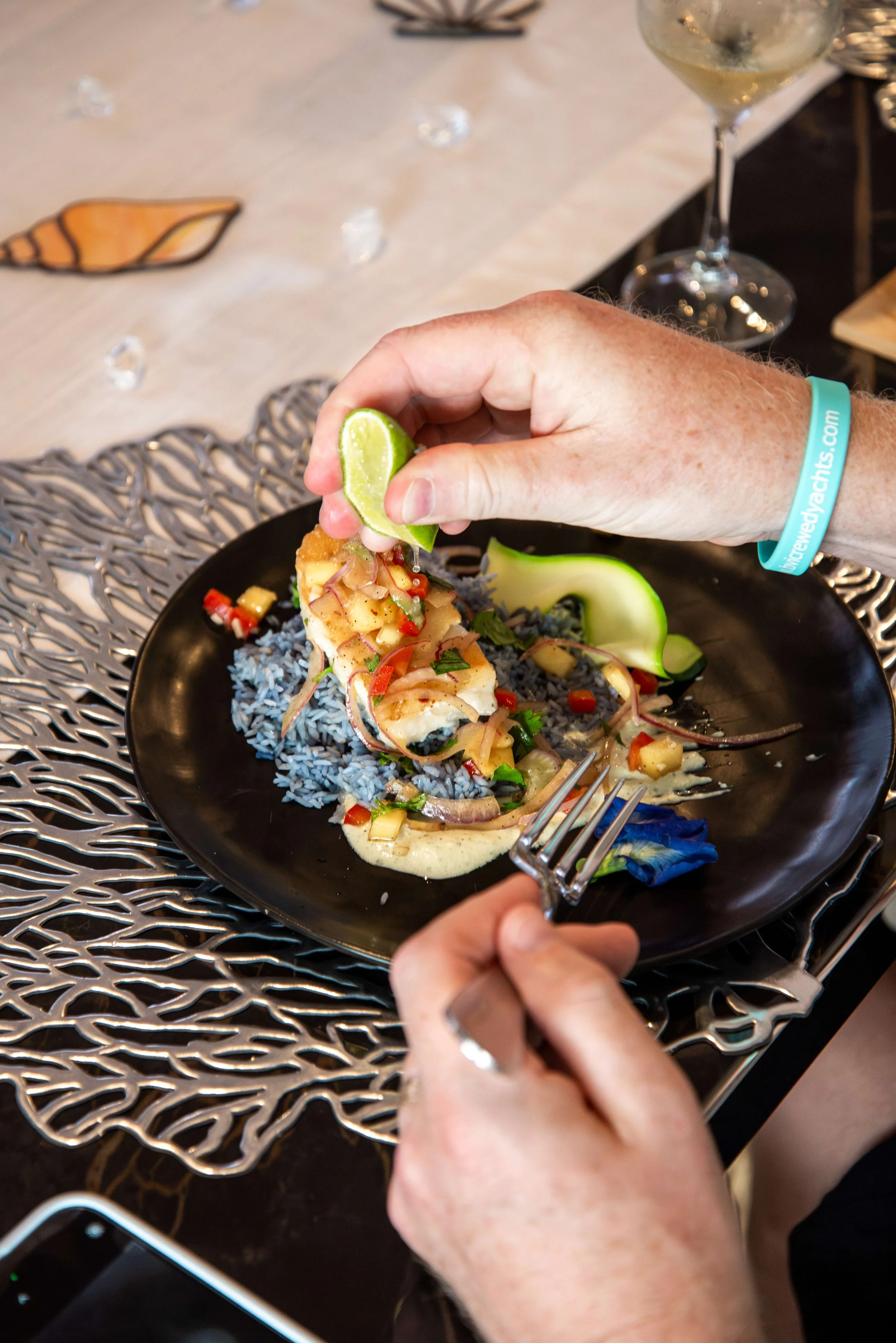 A person squeezing lime over a savory dish of fish topped with diced vegetables and herbs, served on a black plate with blue rice and garnished with a cucumber slice, on a decorative metal placemat.