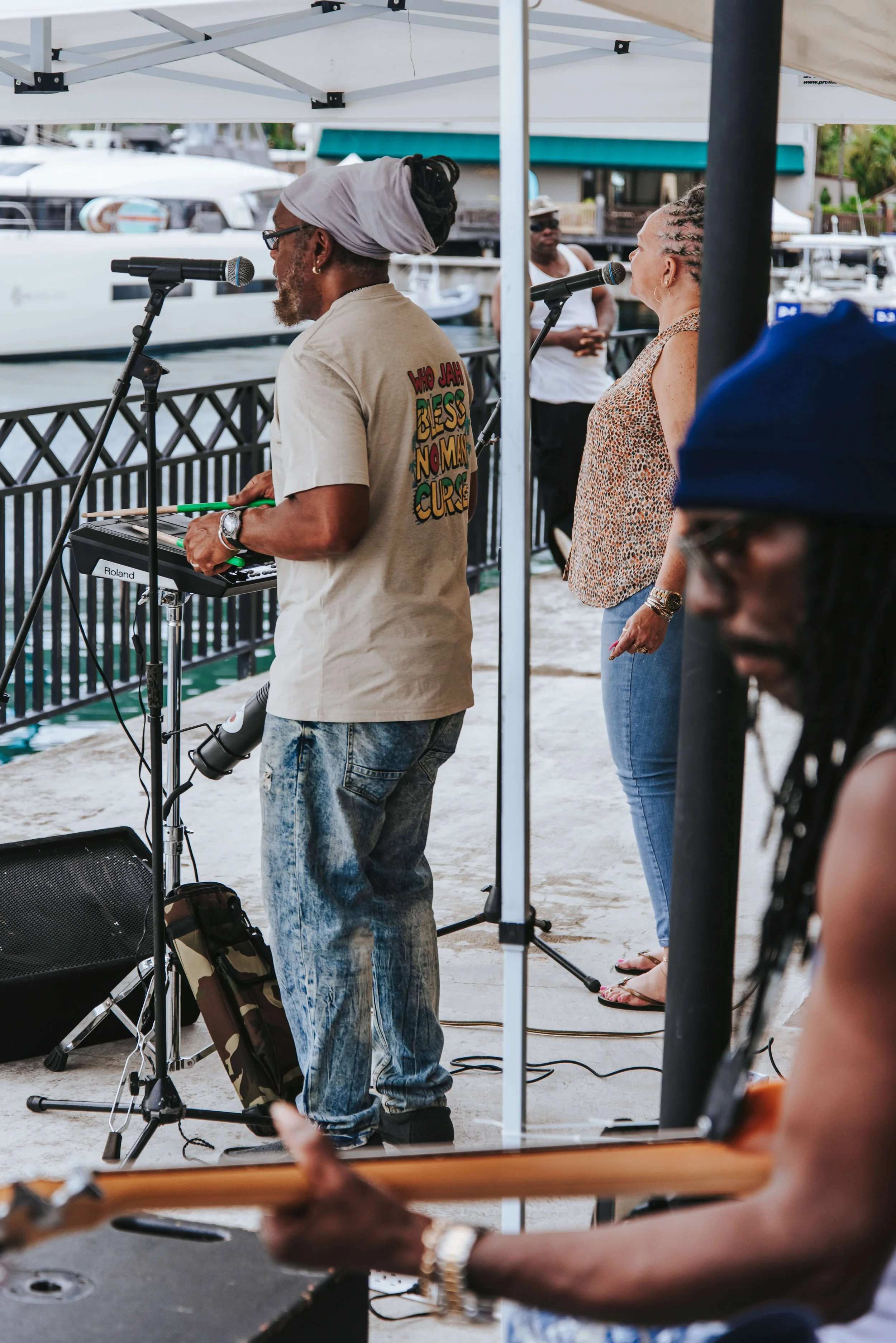 Musicians performing outdoors near a marina with boats in the background, under a canopy, including a man with dreadlocks playing guitar and a woman singing.