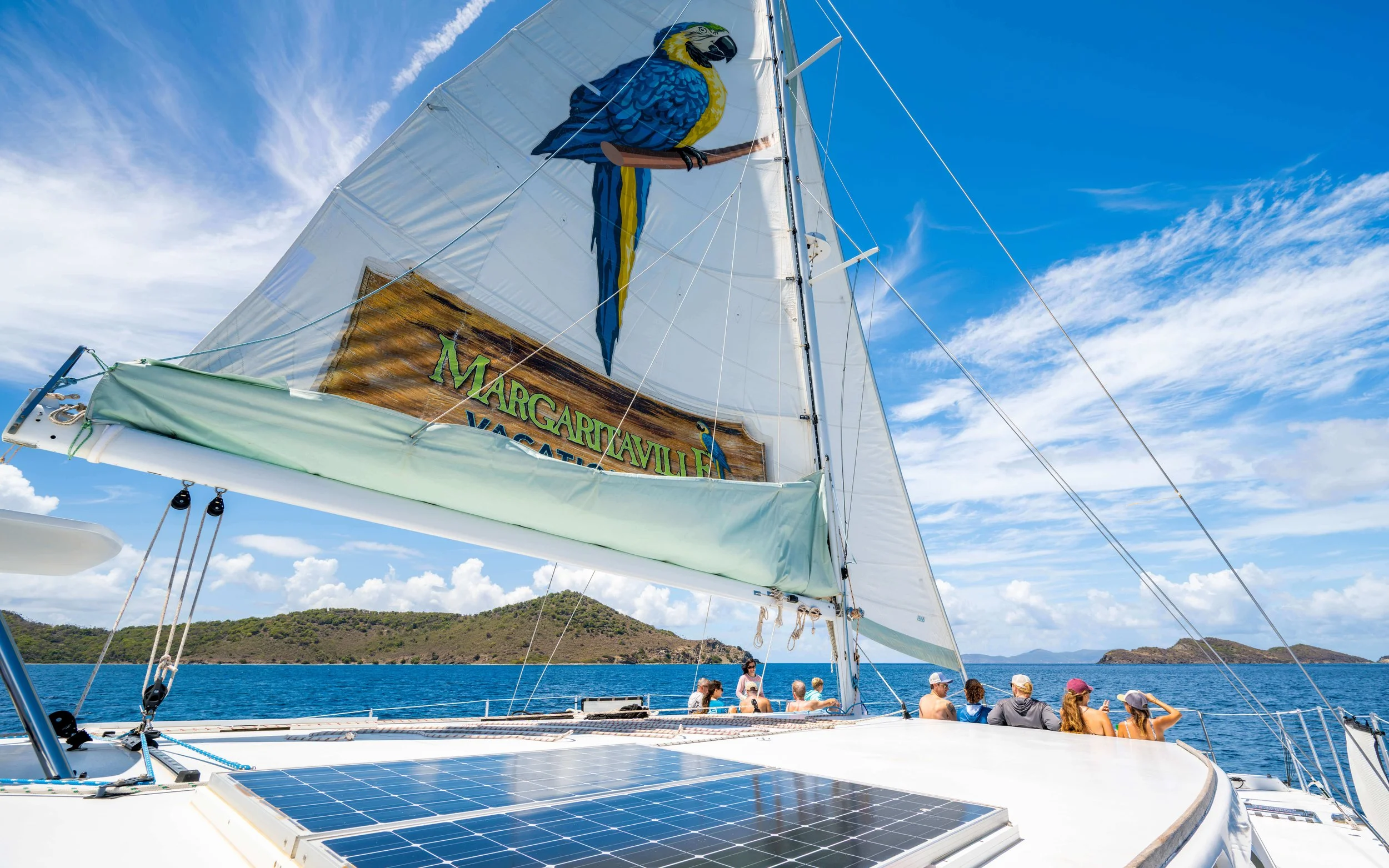 A sailboat with a white sail displaying a colorful parrot graphic, on a body of water with a hilly landscape in the background. Several people are on the boat, some wearing hats, enjoying the sunny day.