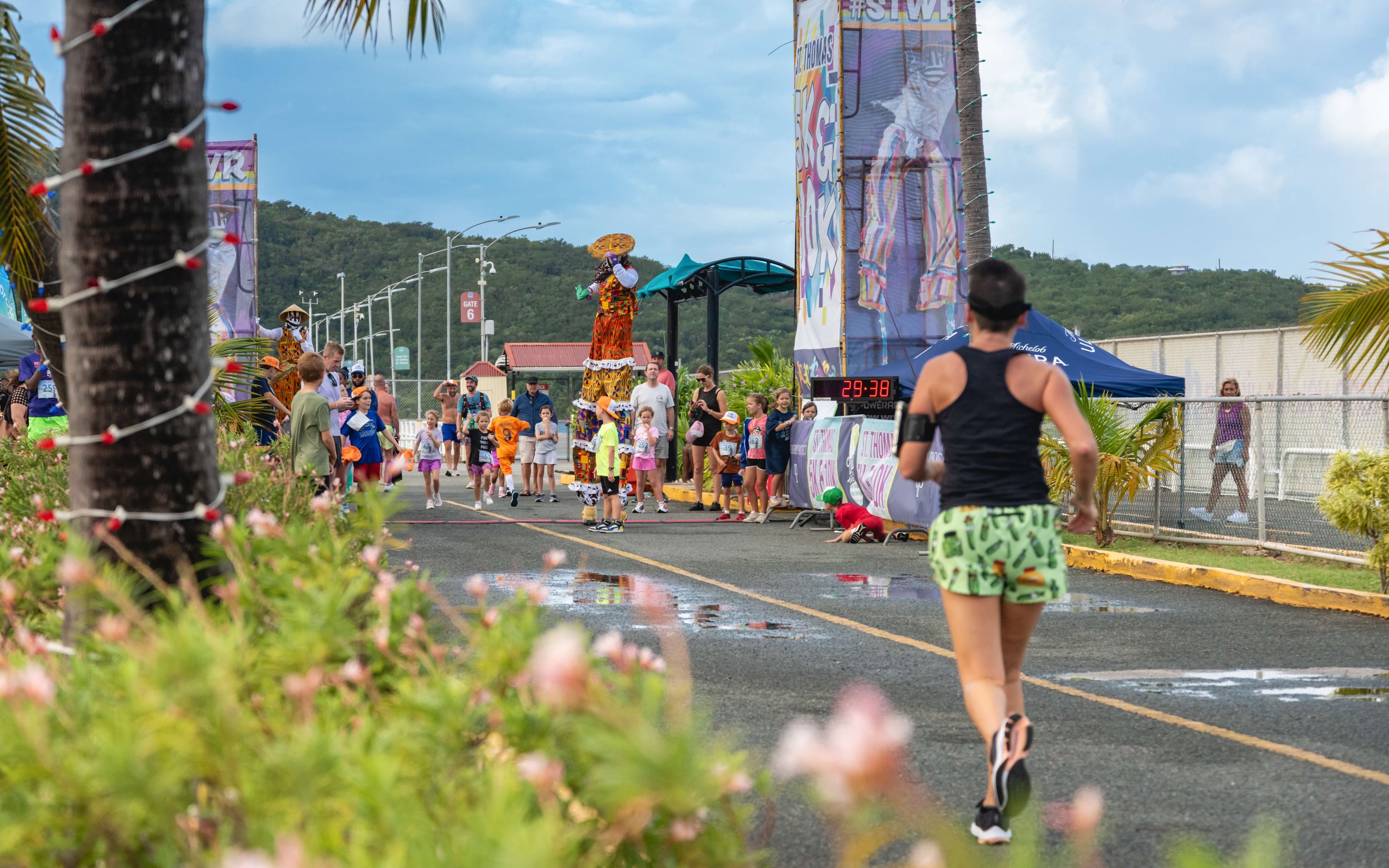 Person in black tank top and colorful shorts running in a festive outdoor race event with children and adults, colorful decorations, and large digital clock showing 29:38.