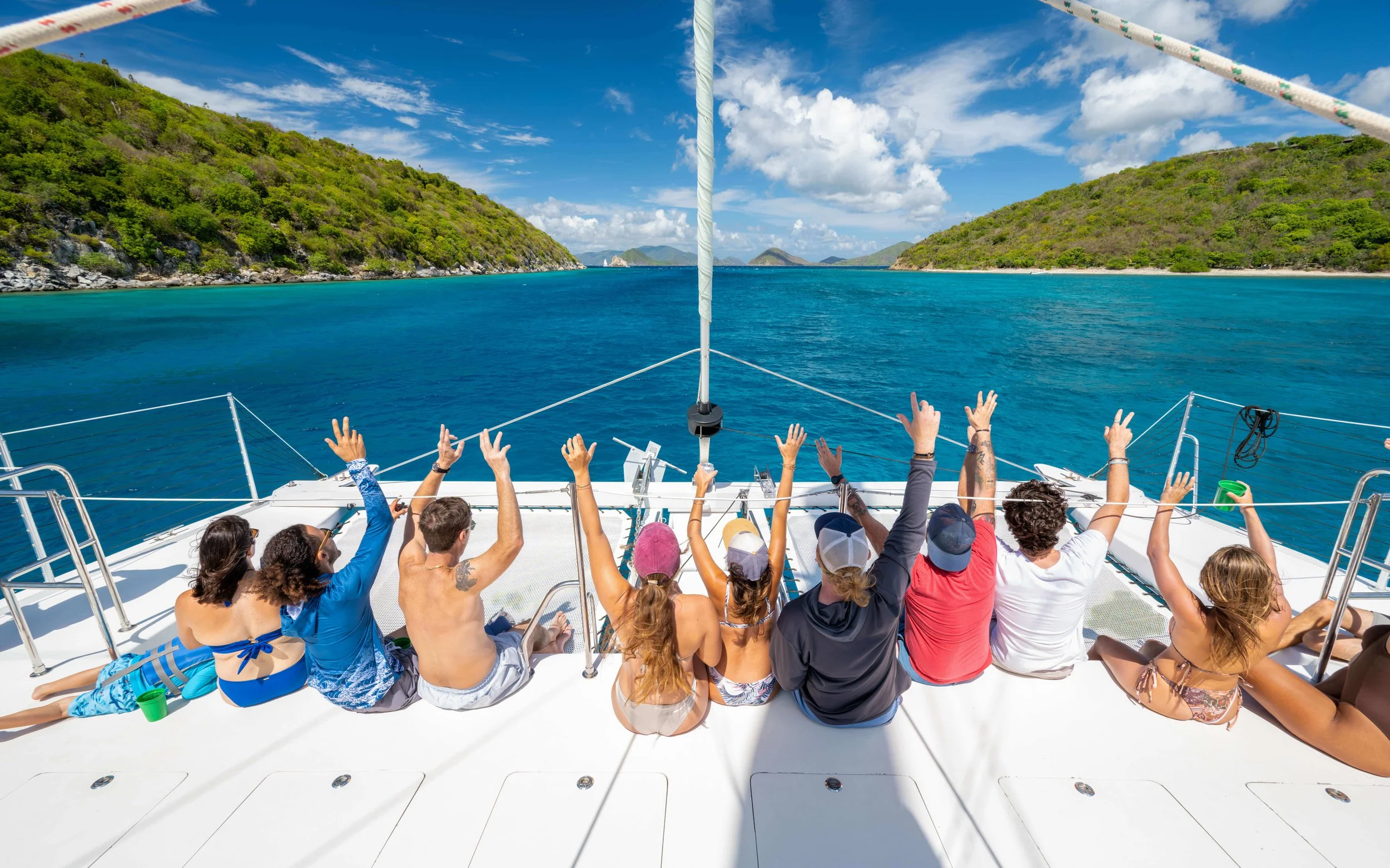People sitting and raising their hands on a boat with a scenic ocean view and islands in the background under a partly cloudy sky.