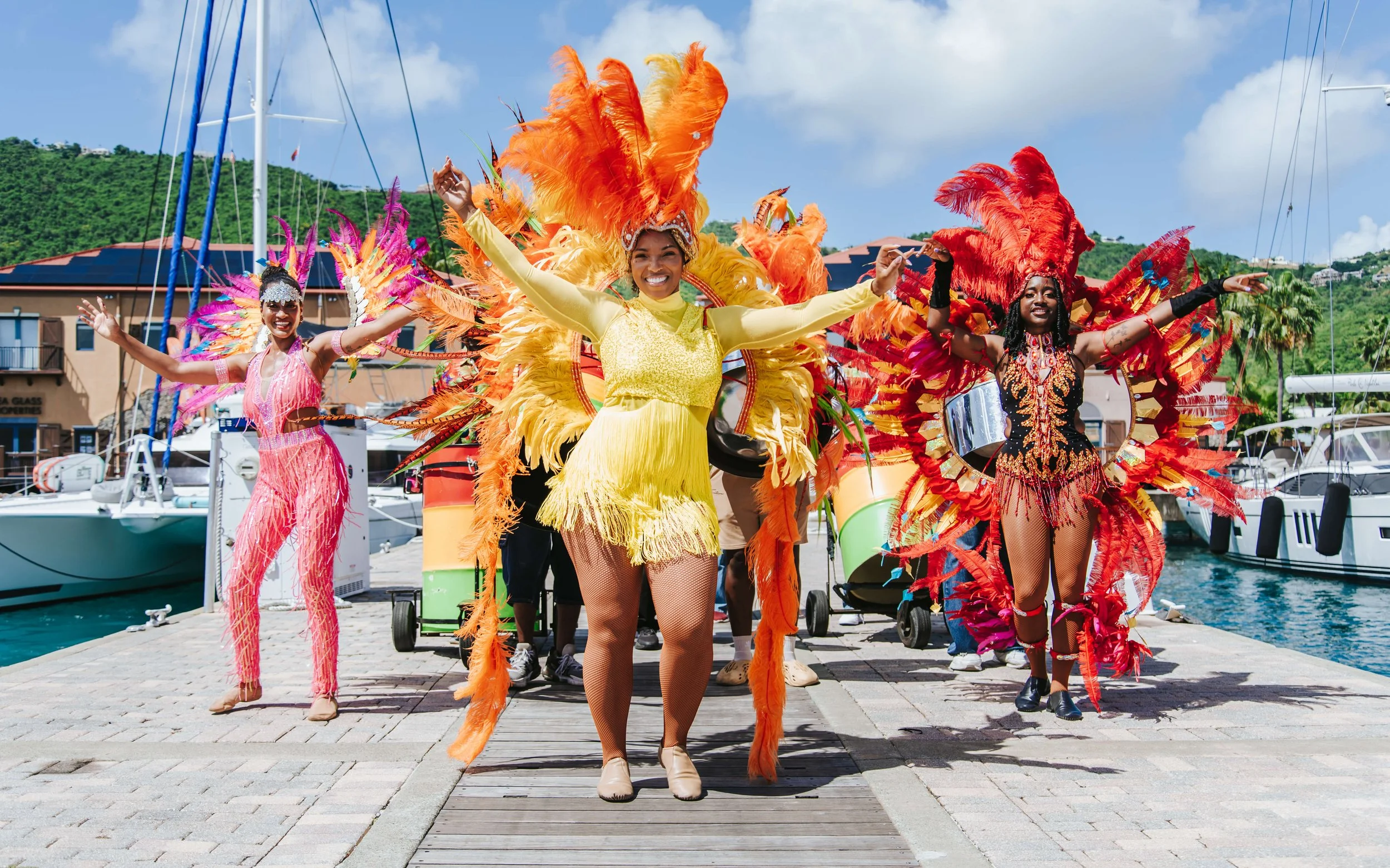 Group of people in colorful carnival costumes dancing on a dock with boats, hills, and blue sky in the background.