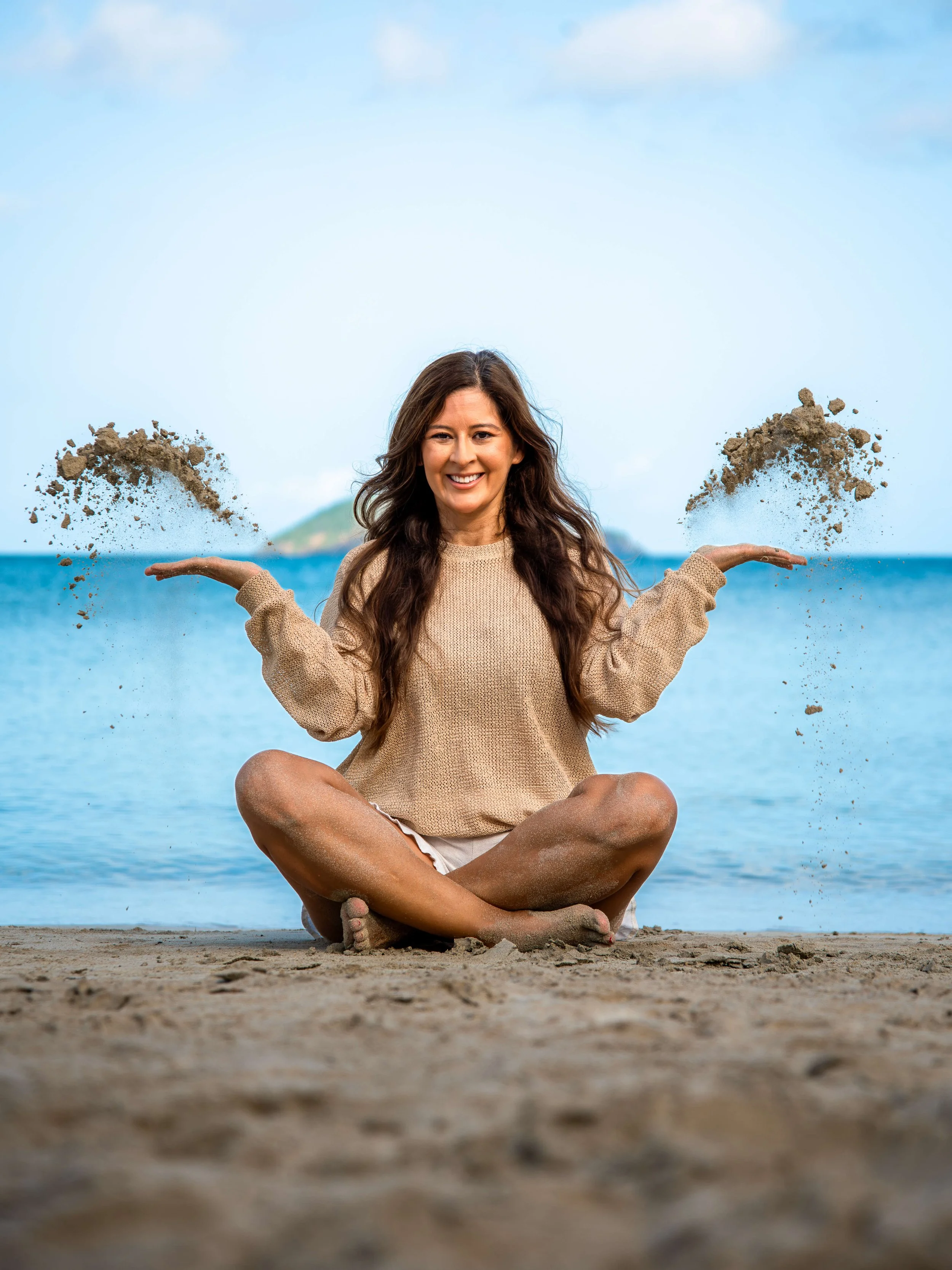 Woman sitting cross-legged on the beach, smiling, with sand falling from her hands near the water and sky in the background.