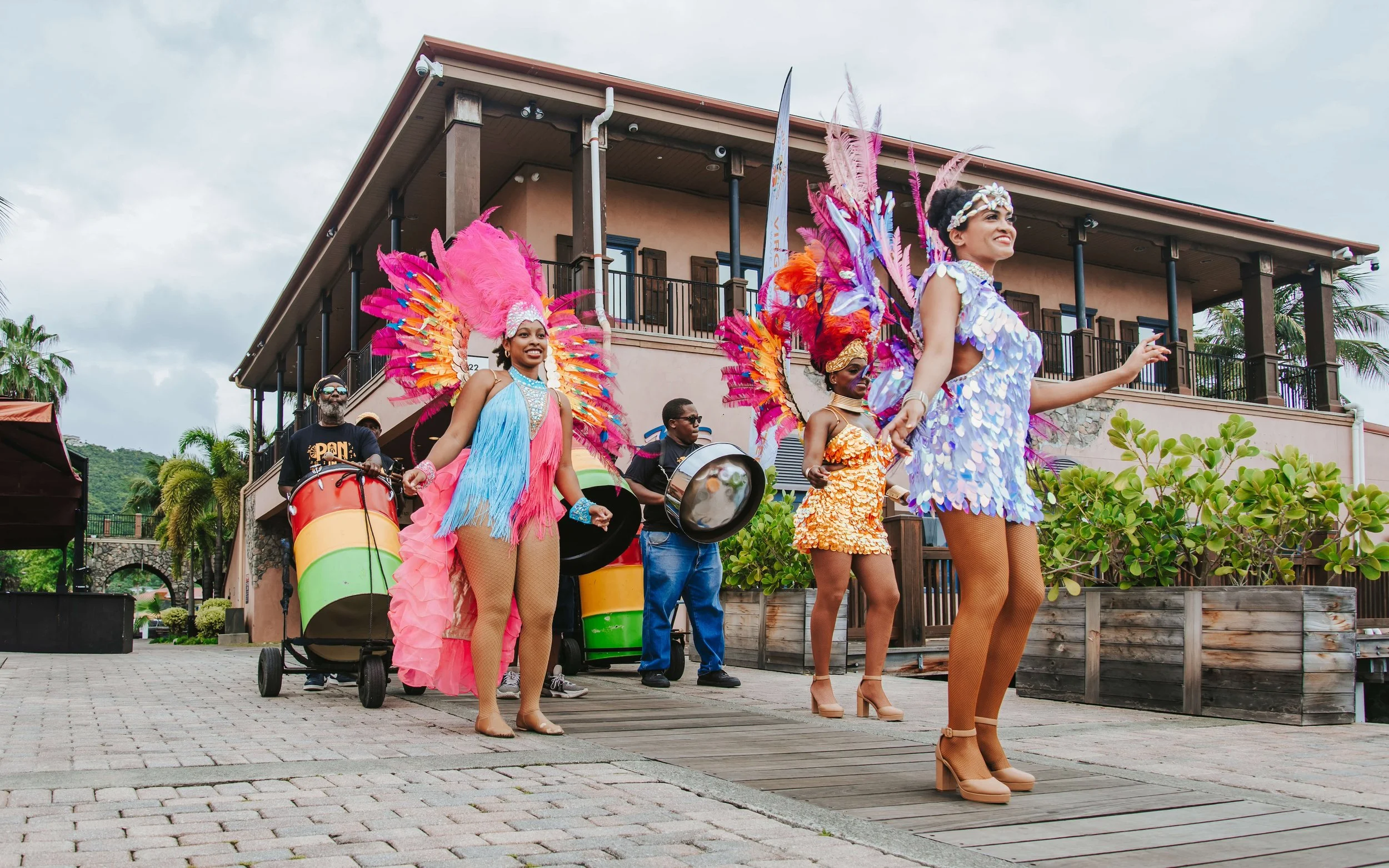 A colorful parade with women in vibrant costumes with feathers, playing musical instruments, walking on a wooden and brick pathway outside a building with palm trees.