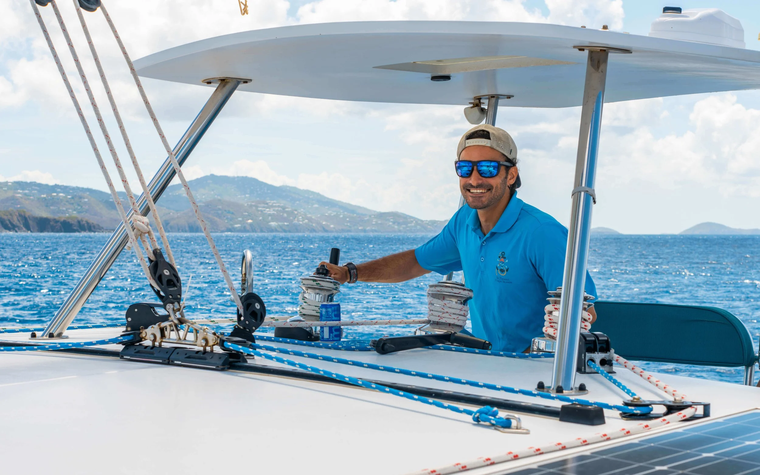 A man with a beard, sunglasses, and a cap smiling and steering a sailboat, with a scenic view of the ocean and hills in the background.