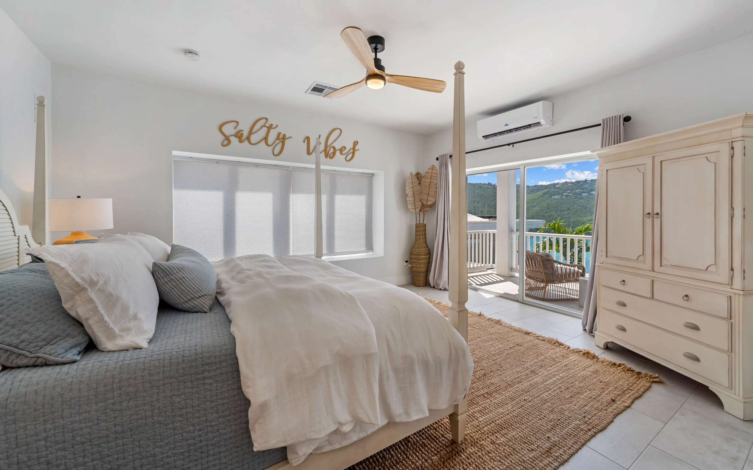Bright bedroom with white bedding, a wooden canopy bed, and a view of mountains outside through sliding glass doors, decorated with coastal-themed accessories and a 'Salty Vibes' sign.
