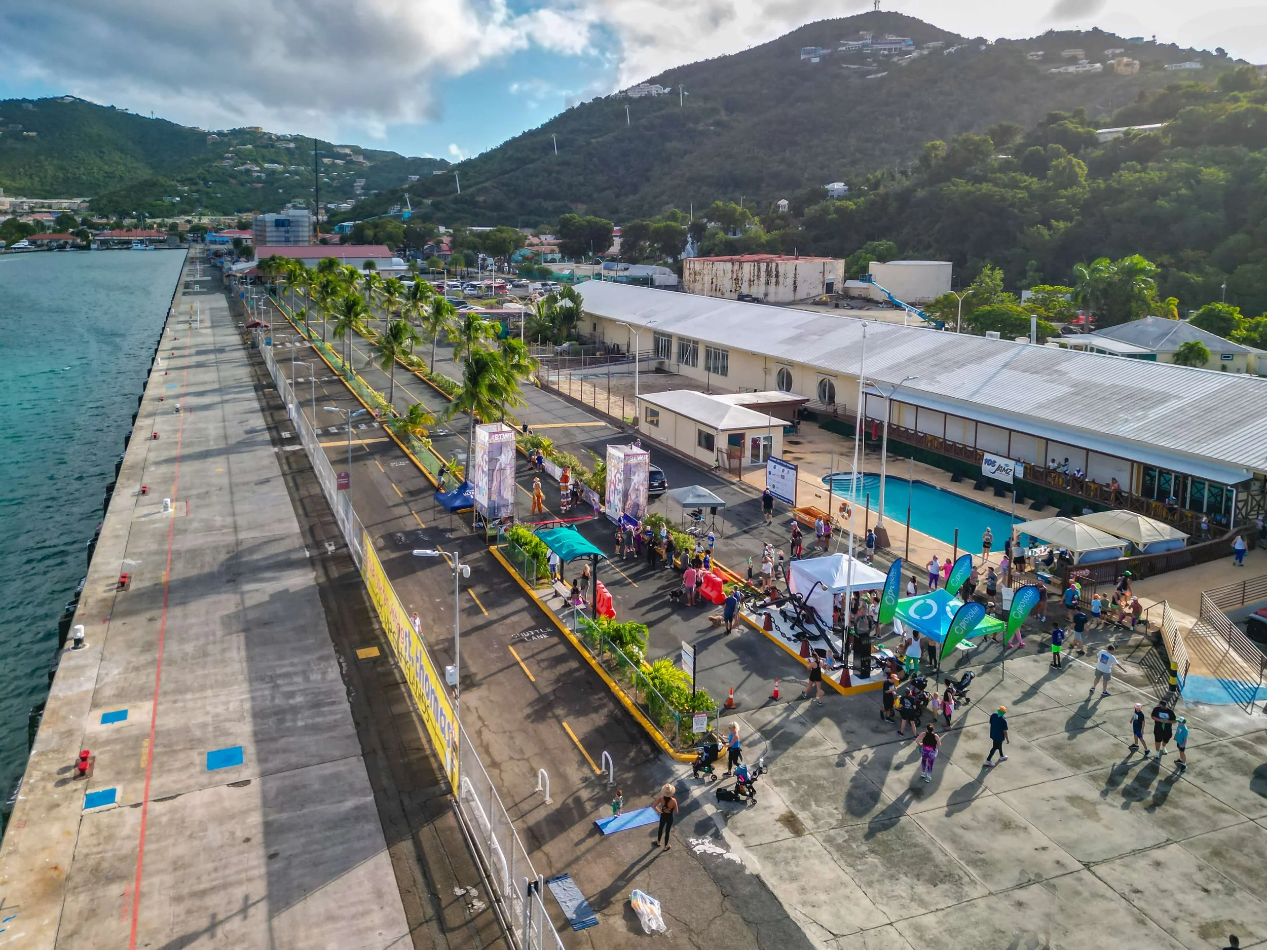 Aerial view of a coastal event with a crowd of people, tents, and booths next to a swimming pool, near a building with metal roofing, with mountains and trees in the background.