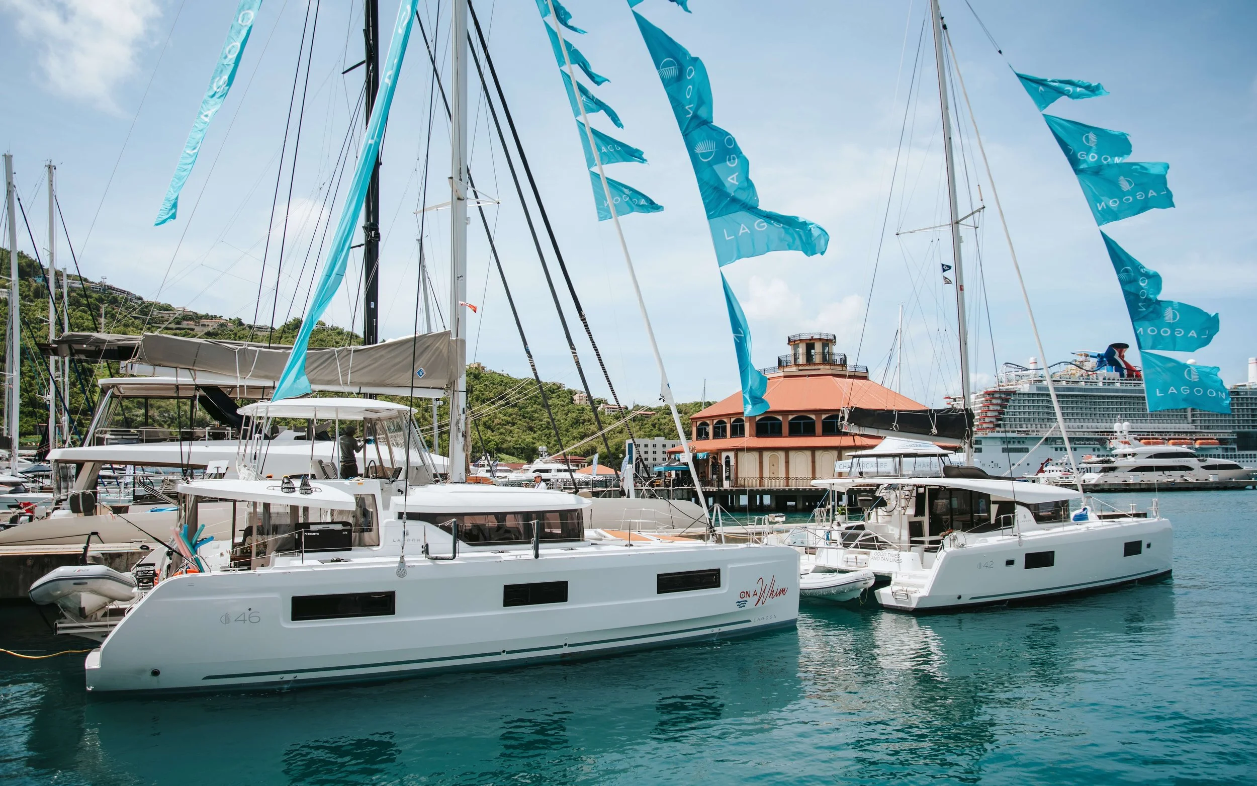 Marina with multiple sailboats and yachts docked, covered in blue flags, near a waterfront building with red roof and a large cruise ship in the background, on a clear day.